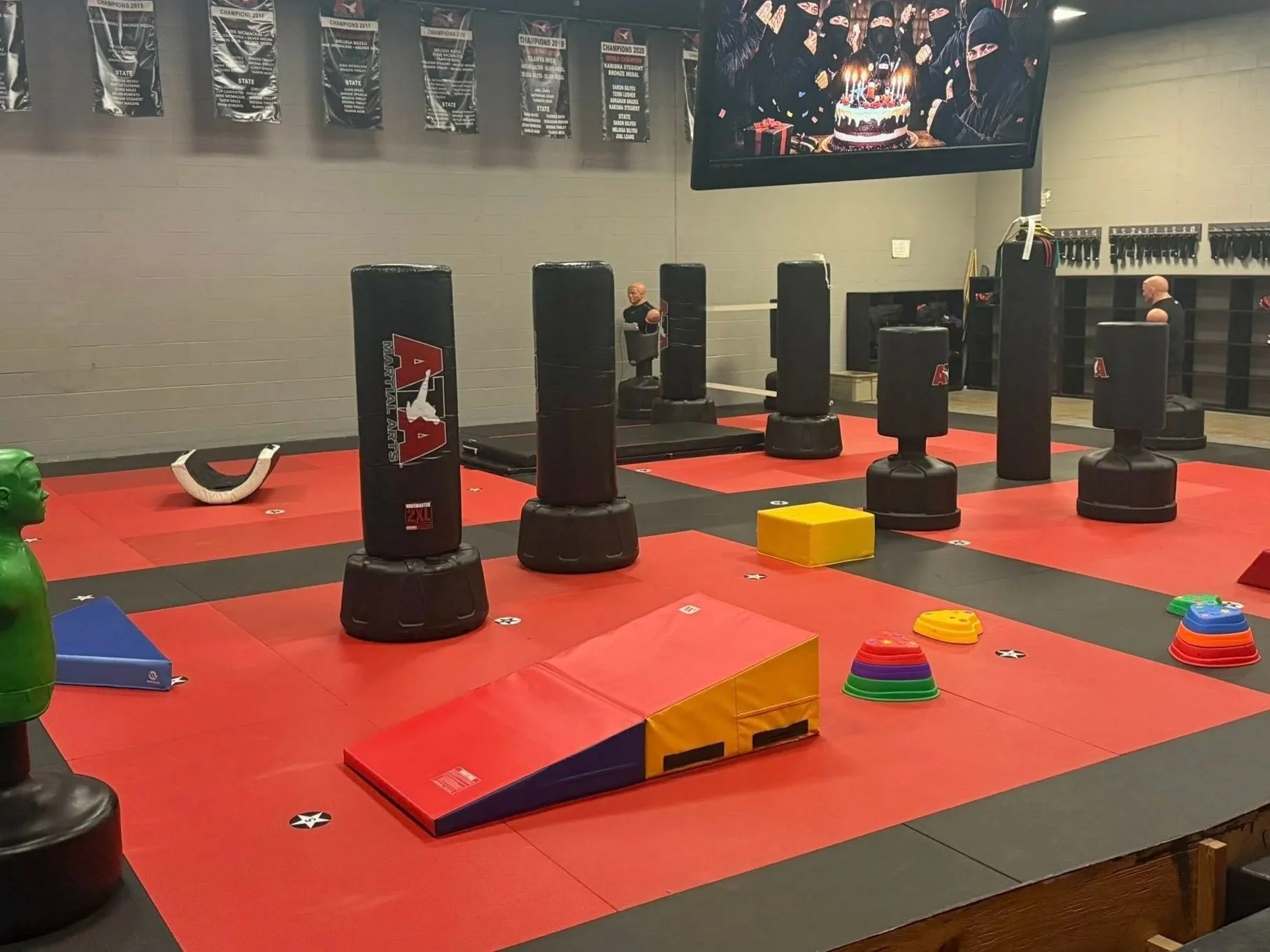 Martial arts training area with punching bags, soft mats, and colorful stepping platforms, in a gym with banners and a large screen on the wall.