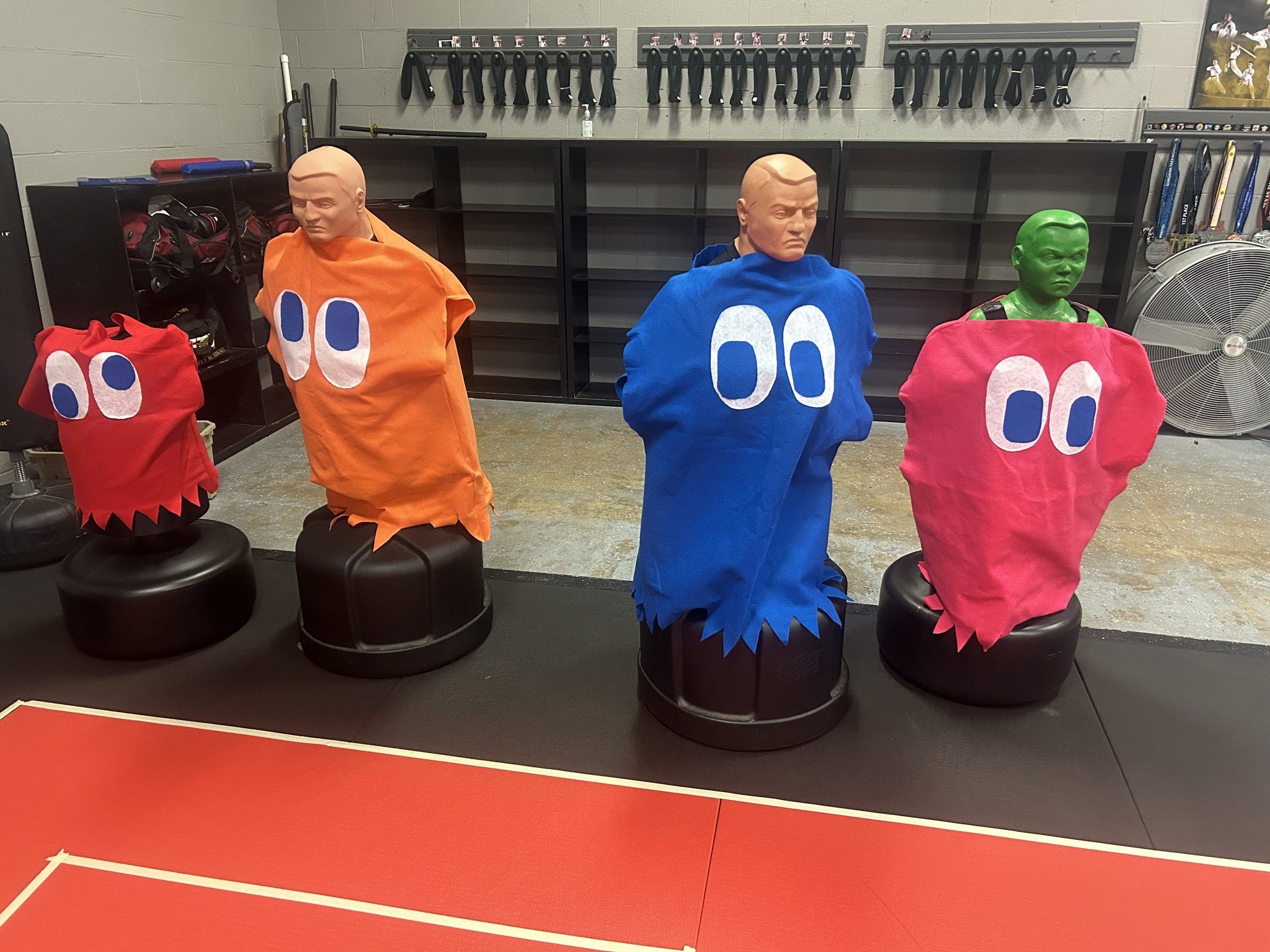 Three martial arts training dummies dressed as Pac-Man characters with foam costumes and painted faces, in a gym with martial arts equipment and a red mat.