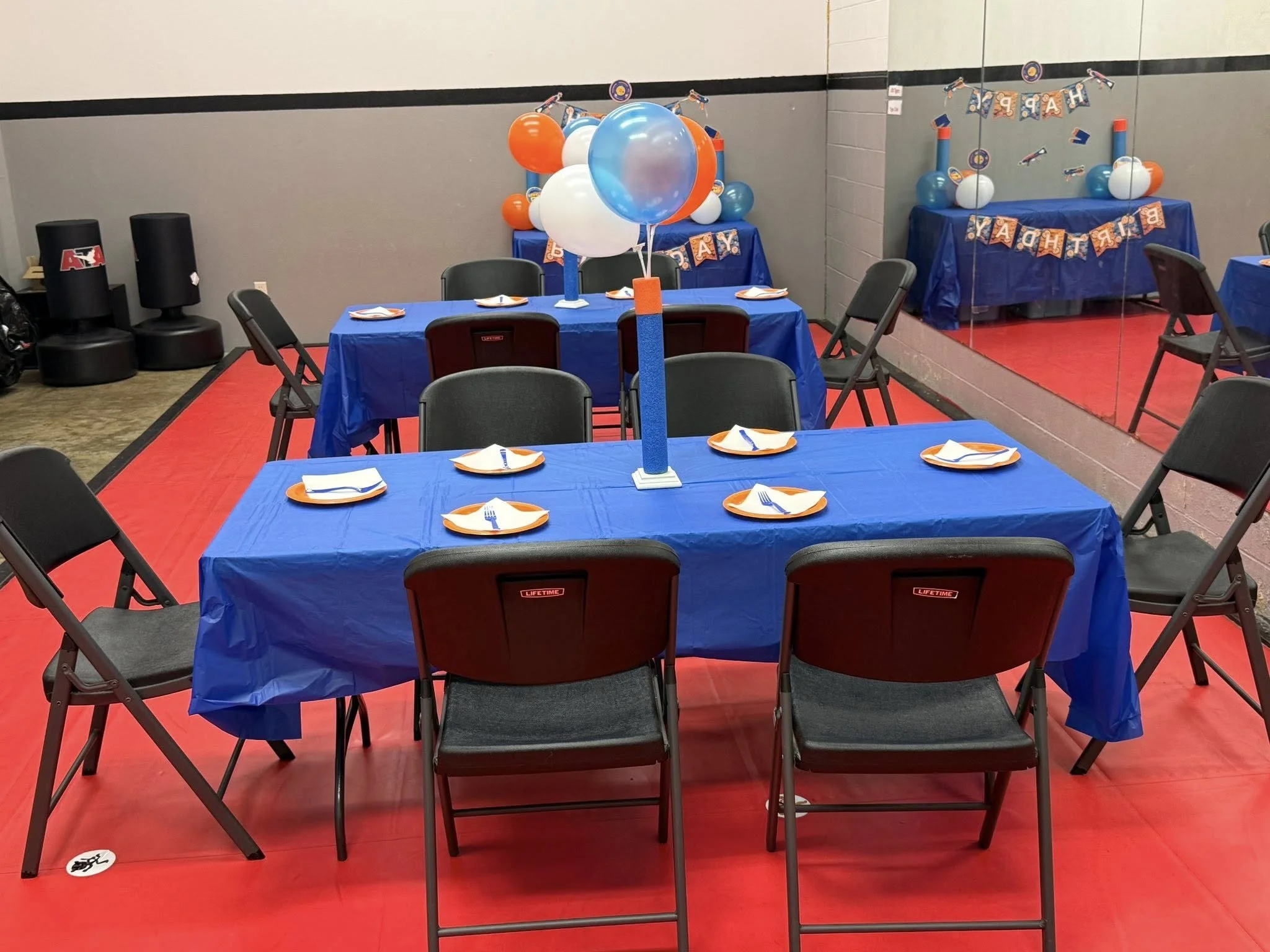 Party setup with three tables draped in blue tablecloths, decorated with balloons and themed banners, with plates, napkins, and utensils arranged on each table.