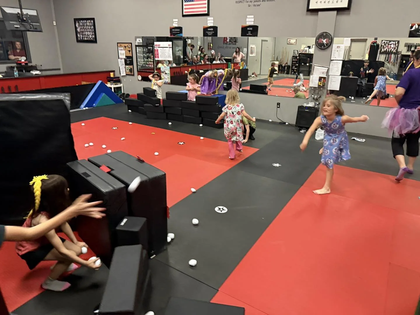 Children playing and running around in an indoor trampoline park with black and red mats, foam blocks, and scattered white balls; adults supervising in the background.
