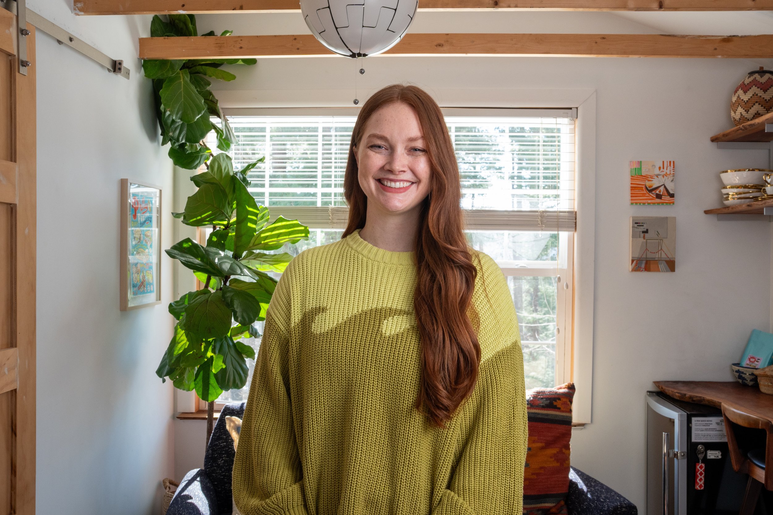 A young woman with long, red hair and a broad smile, Dr. Maureen 
Fleming, wearing a green sweater, standing in a bright room with large windows, green plants, and decorative artwork on the walls.