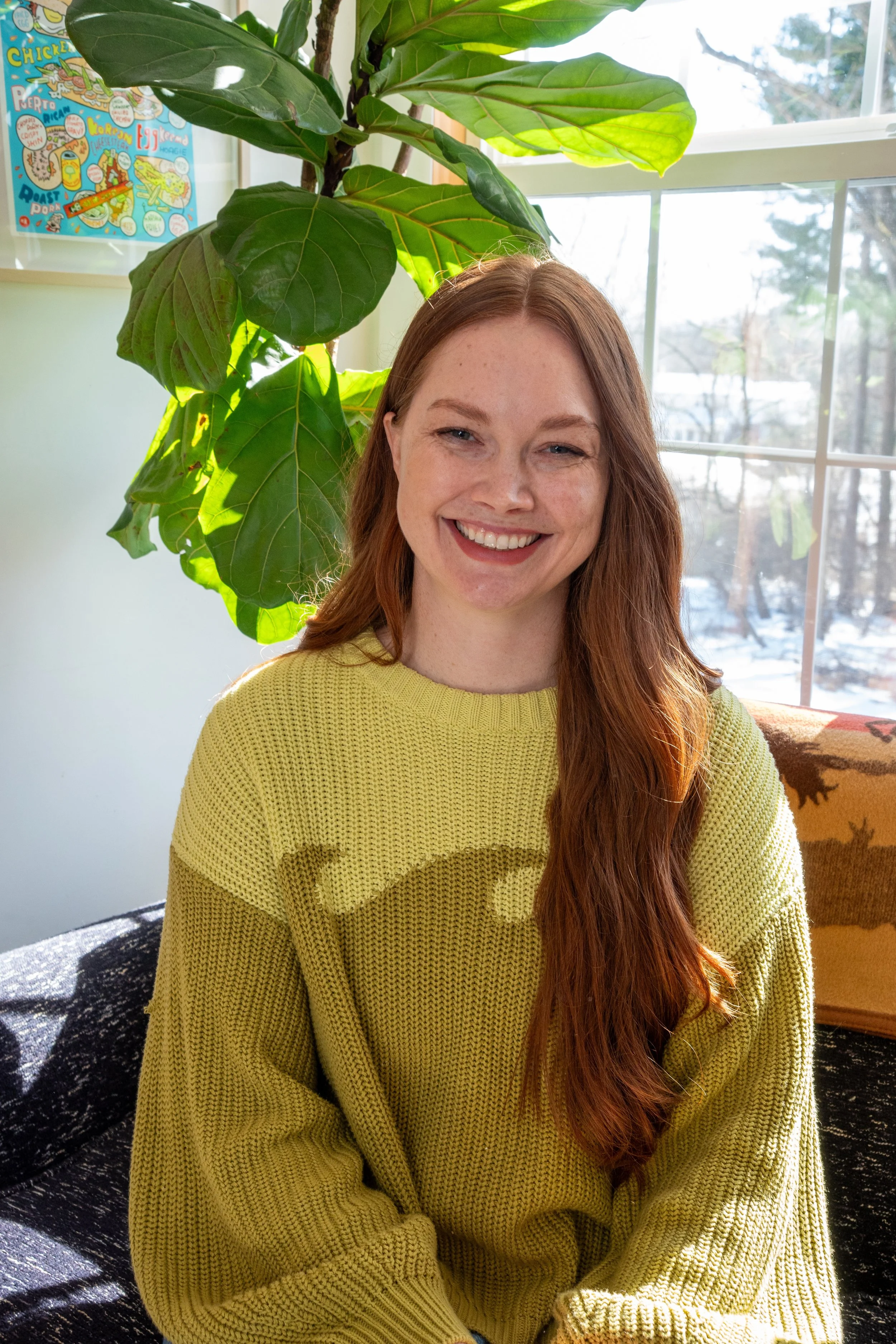 A young woman with long red hair and fair skin smiling while sitting in a sunlit room near a large leafy green plant and a window showing a snowy landscape outside, wearing a yellow knitted sweater.