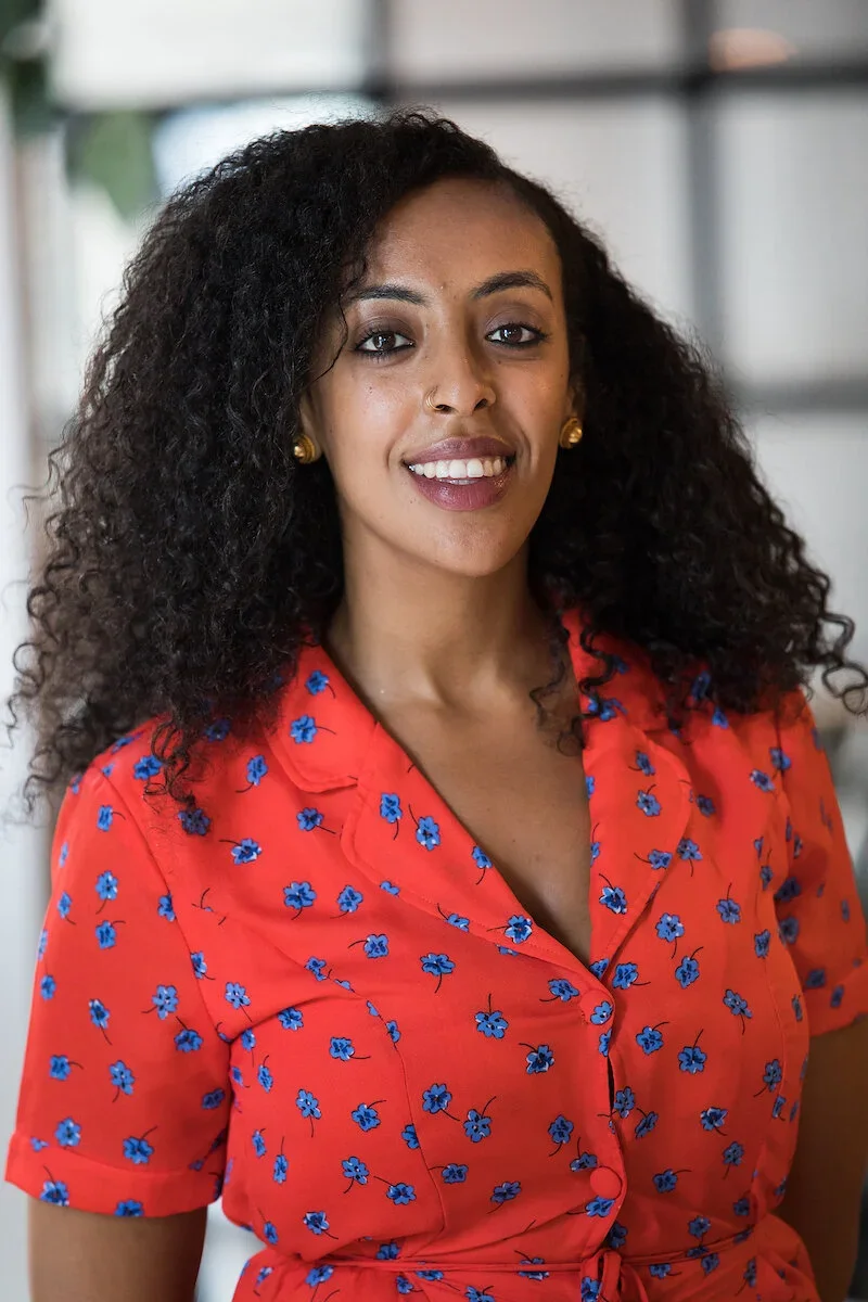 A woman with curly hair, wearing a red dress with blue floral pattern, smiling and looking at the camera.