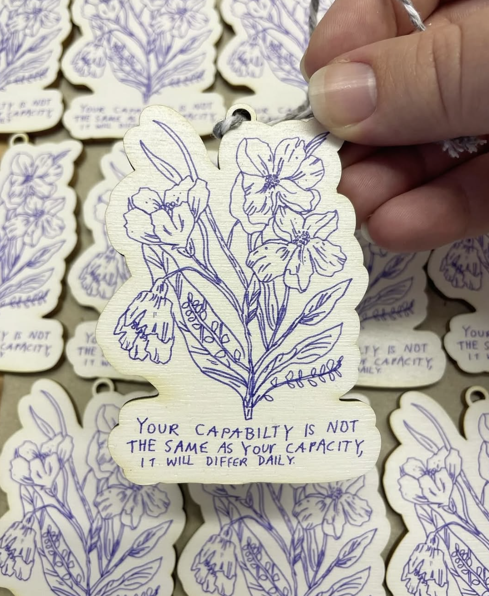A hand holding a wooden tag with a blue illustrated flower design and a motivational quote, surrounded by similar tags.