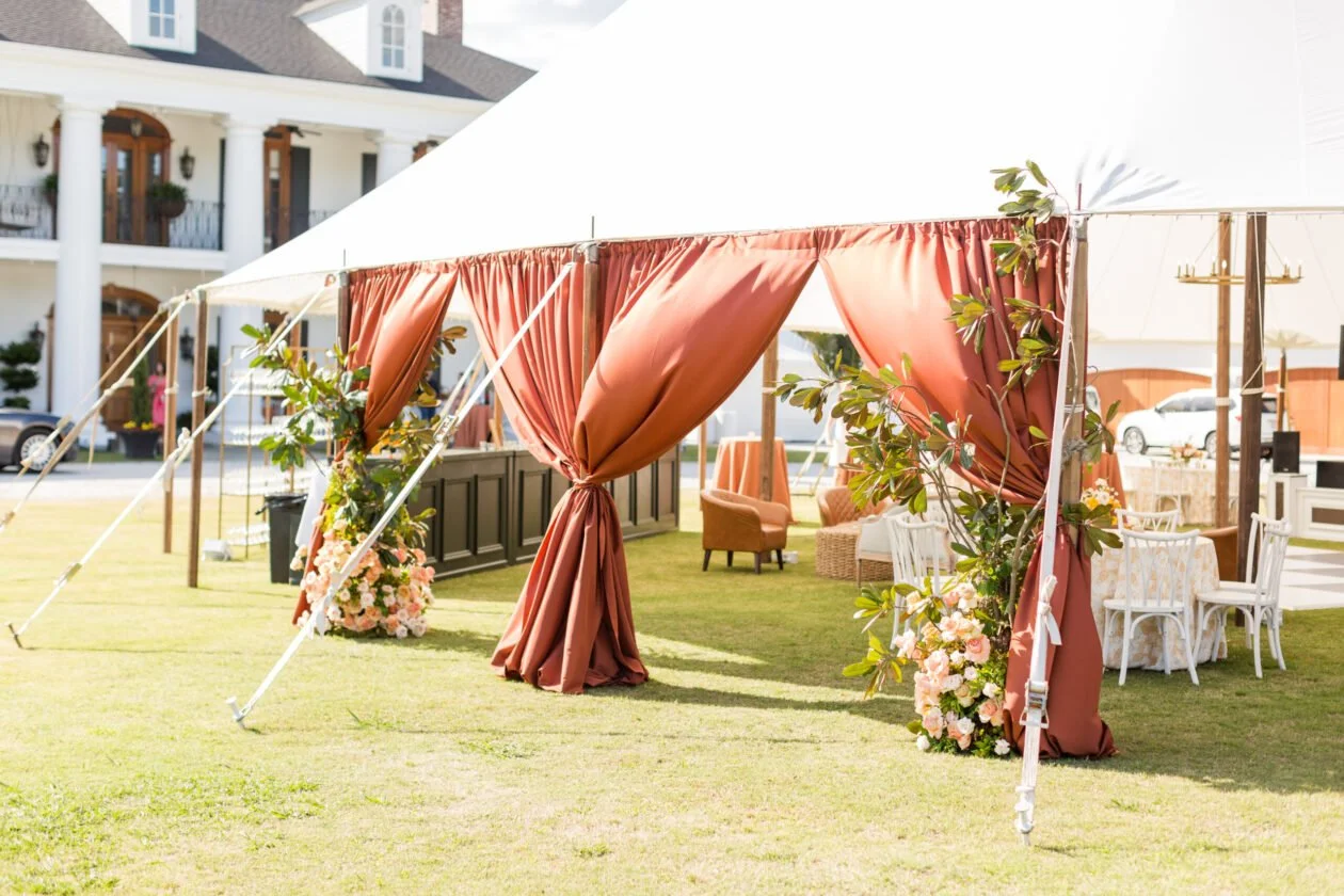 A decorated outdoor tent with rust-colored curtains, floral arrangements, and seating for an event on a lawn in front of a large house.