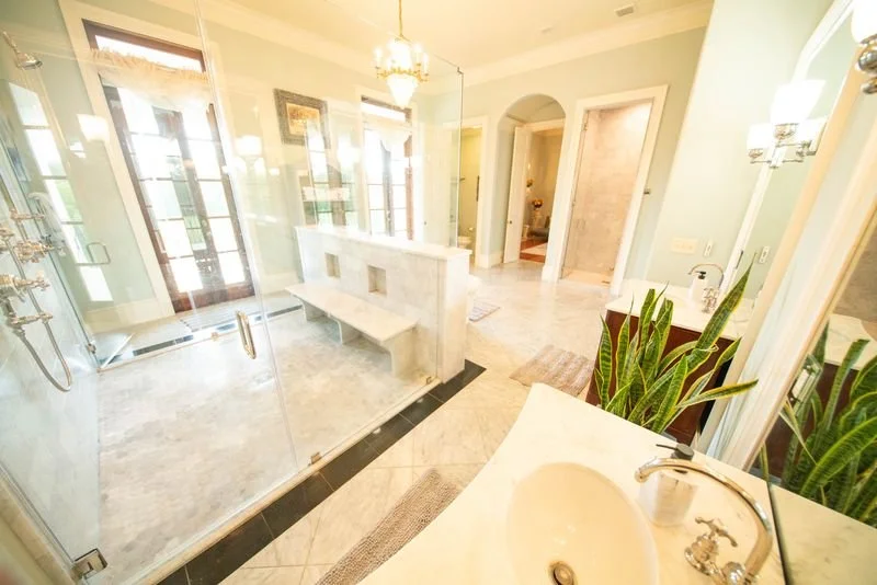 Bright bathroom with glass-enclosed shower, marble surfaces, green plants, and natural light from large windows.