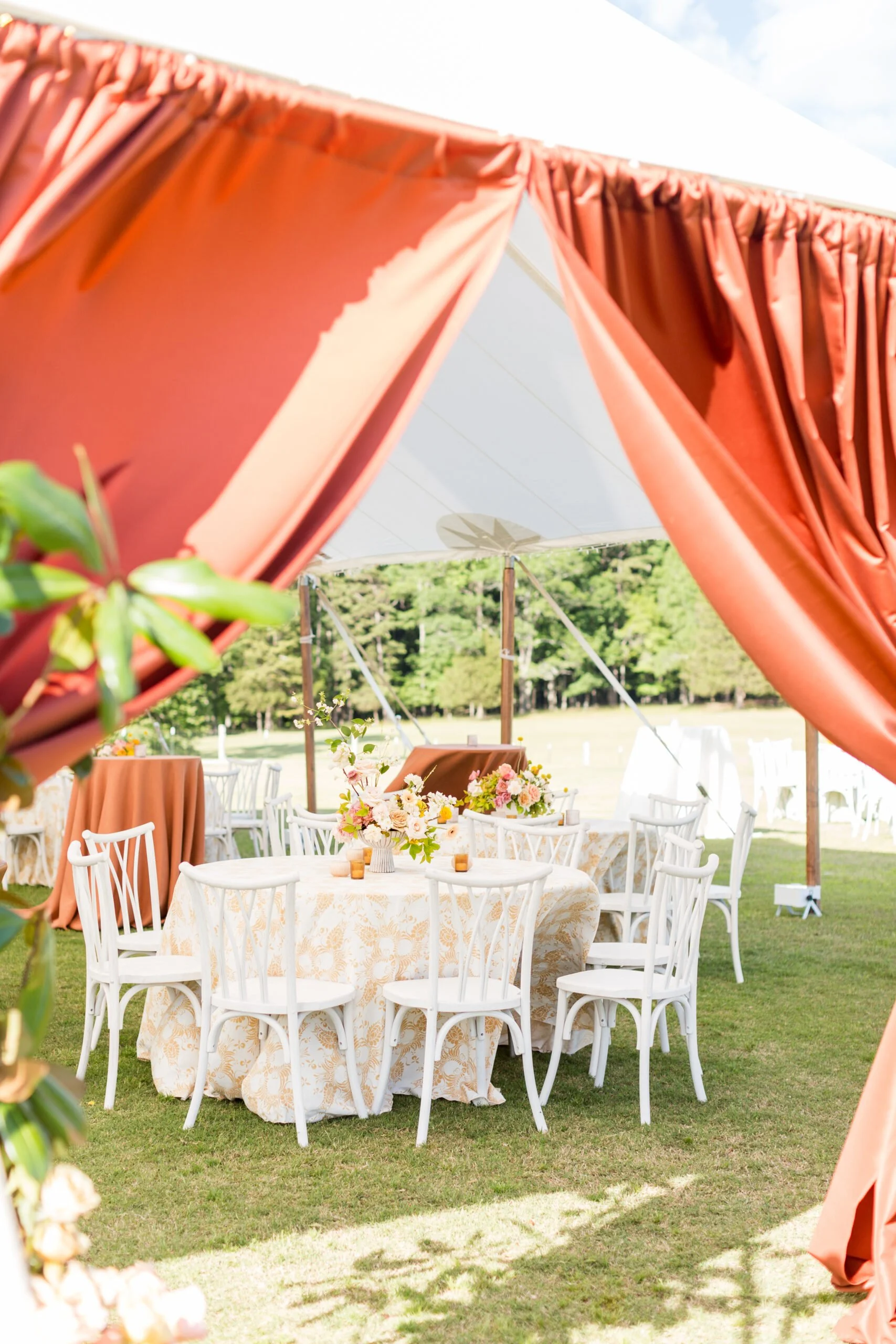Outdoor event setup with a round table, white chairs, floral centerpiece, under a white tent with coral drapes, on grassy ground.
