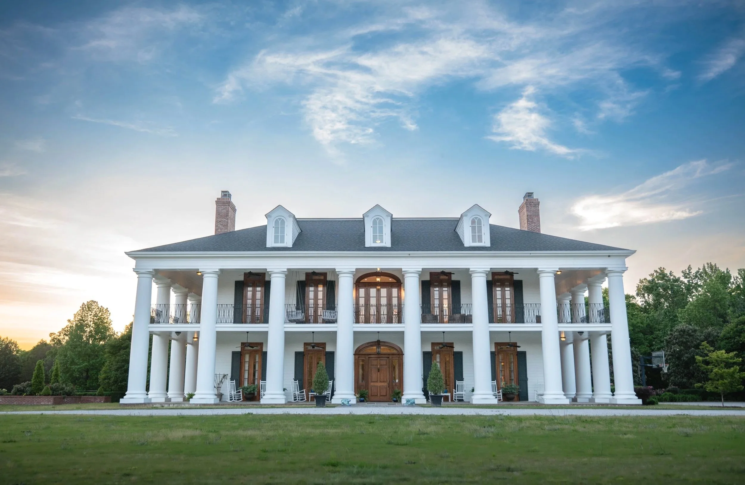 Large white mansion with tall pillars, multiple balcony doors, and a dark roof set against a blue sky with clouds, surrounded by greenery.