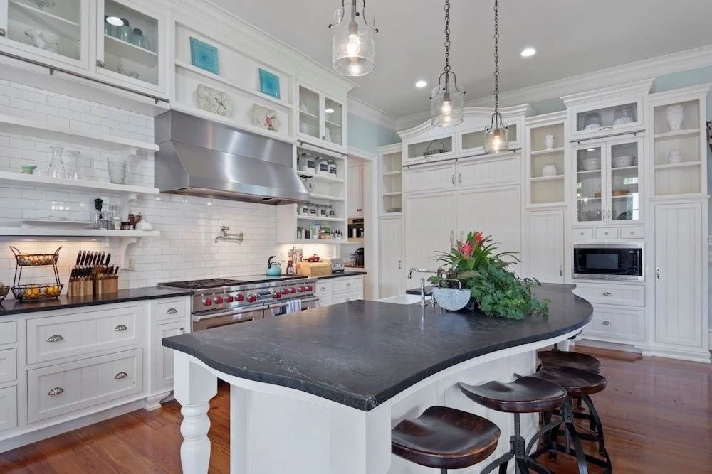 Modern kitchen with white cabinets, a large black countertop island, stainless steel stove and range hood, open shelving, and three pendant lights.