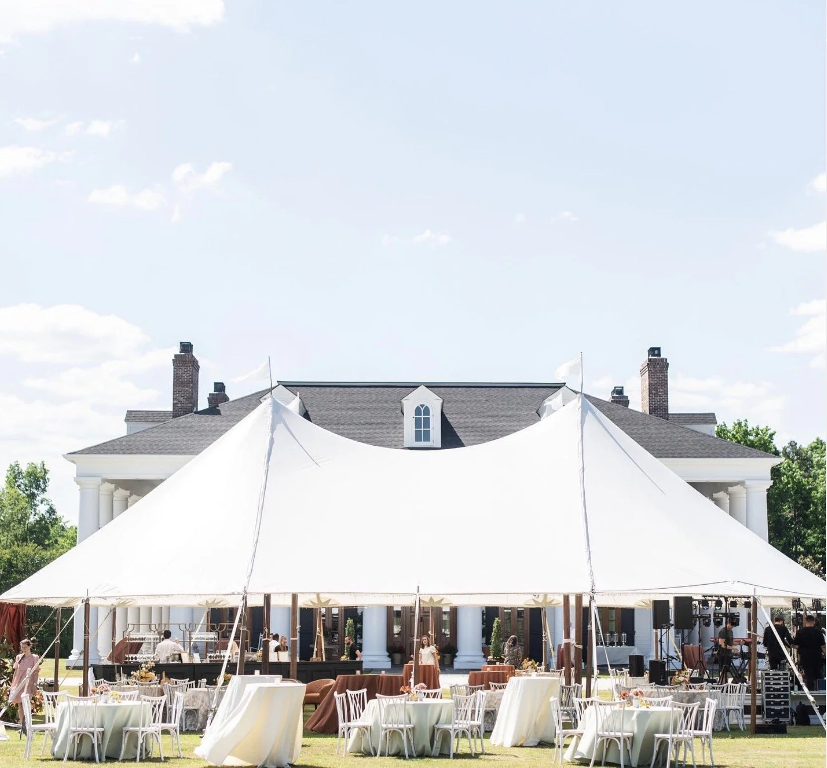 Large white event tent set up on the grass in front of a stately house with columns, surrounded by tables and chairs, with some people preparing for a gathering.