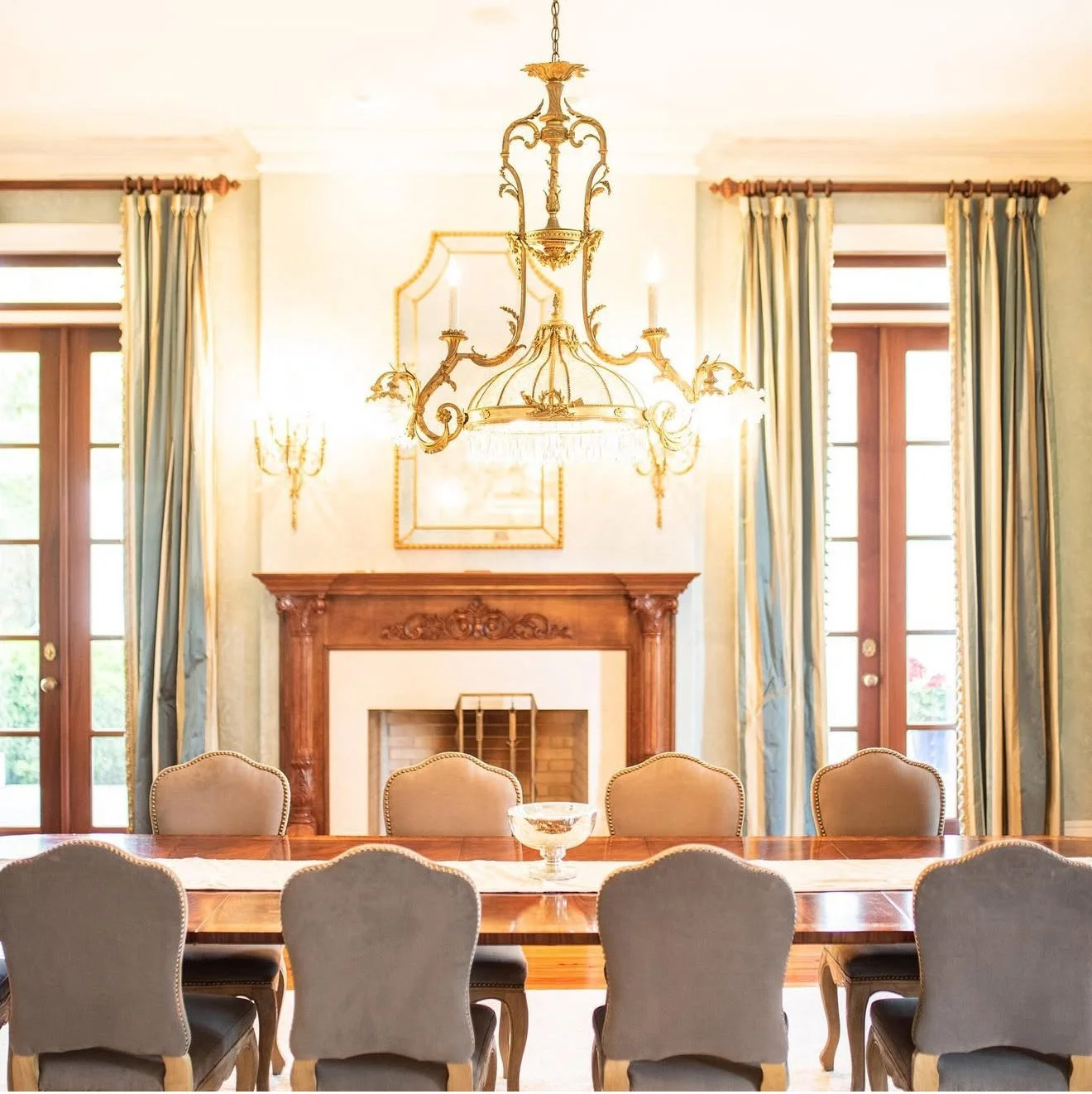 Elegant dining room with a wooden table, upholstered chairs, a carved wooden fireplace, large windows with blue curtains, and a chandelier over the table.