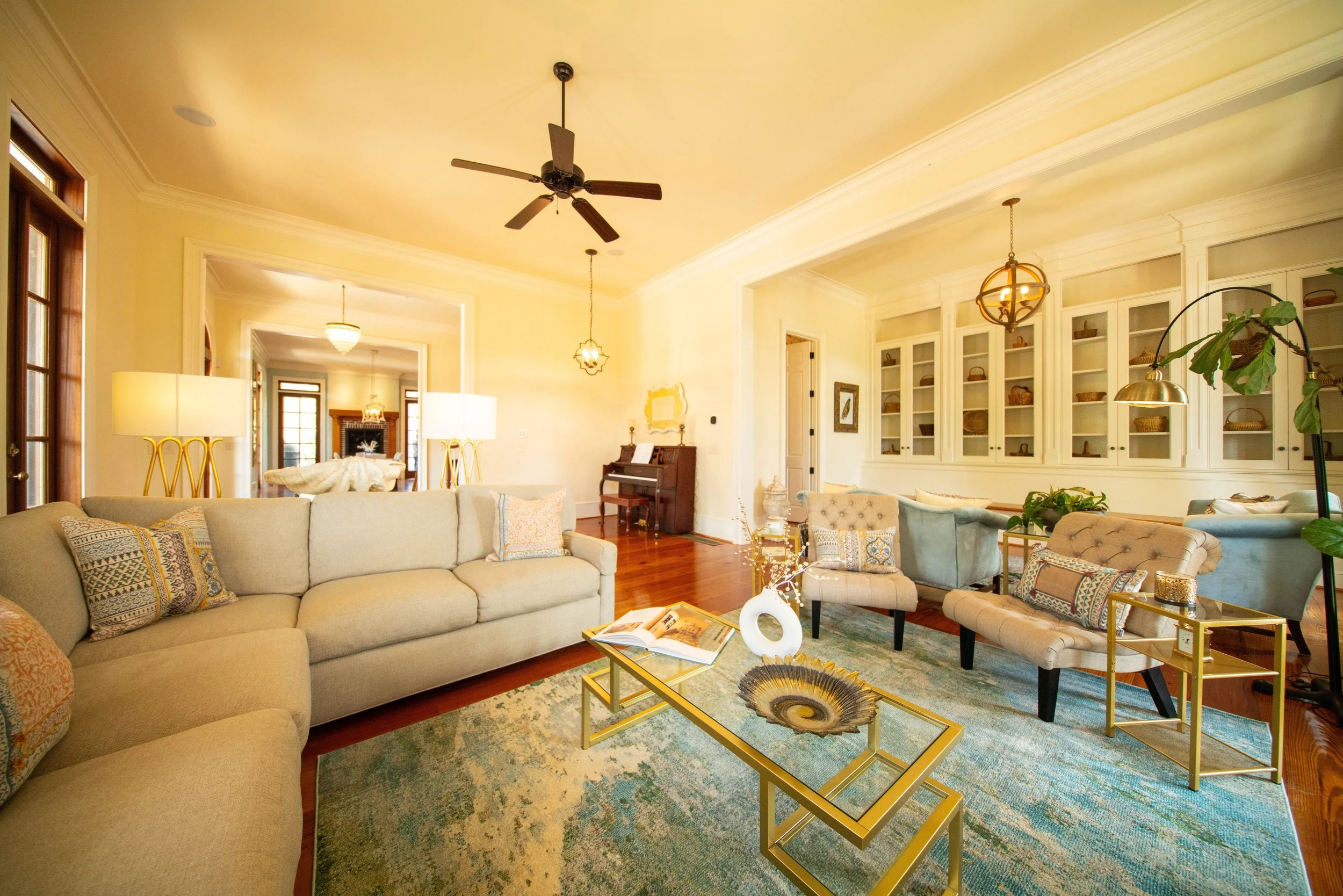 Bright living room with beige sofas, glass coffee table, decorative pillows, large area rug, built-in white cabinets, and various lighting fixtures.