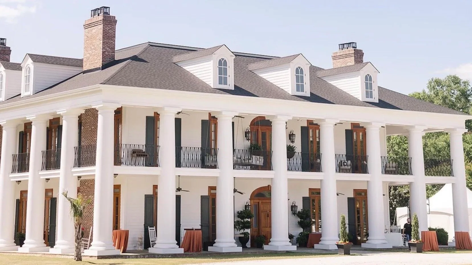 Large two-story white house with tall white columns, brown window frames, and a dark shingle roof, surrounded by green trees and outdoors.