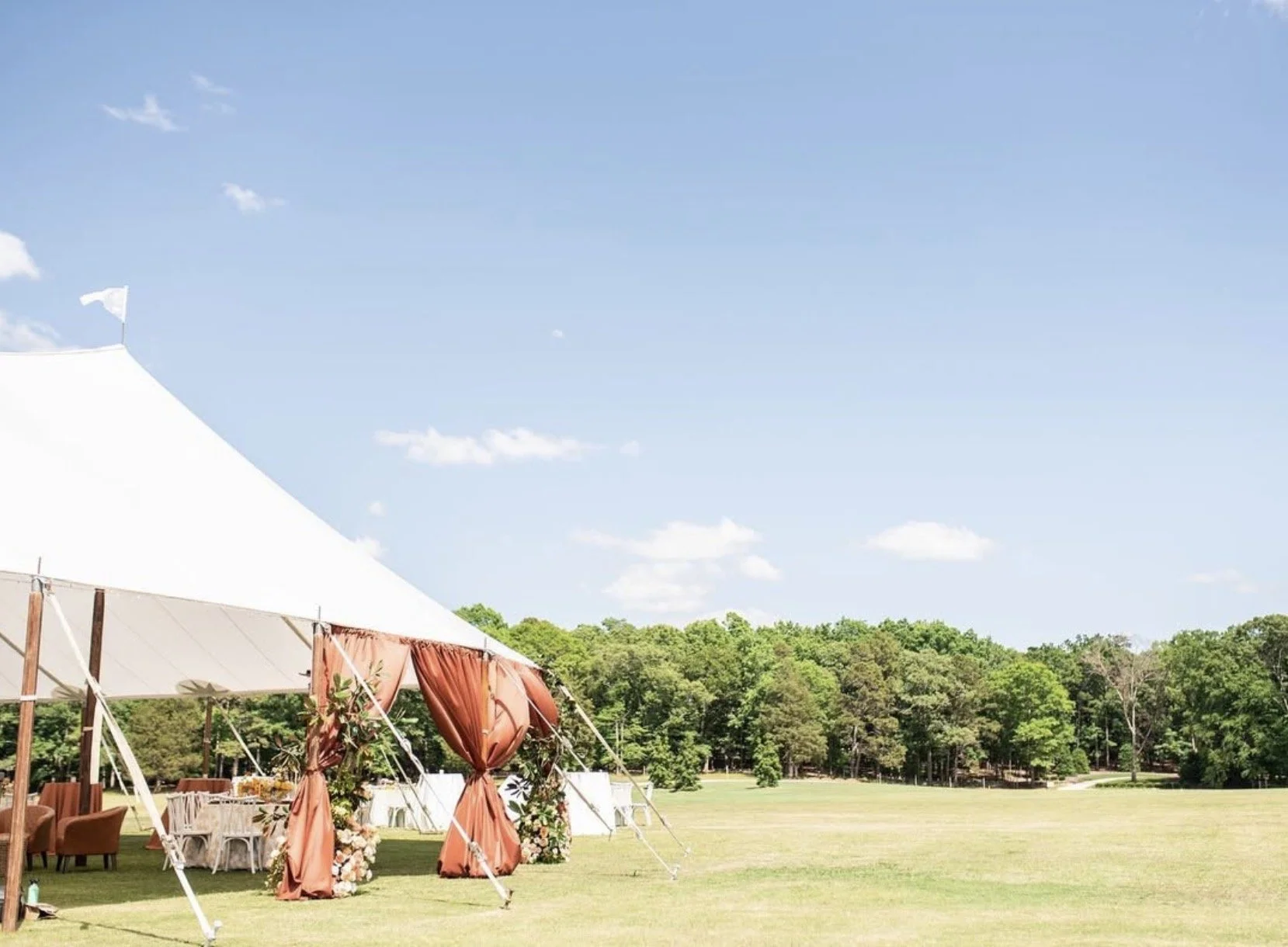 An outdoor wedding setup on a sunny day with a large white tent, decorated with rust-colored curtains and floral arrangements, on a lush green field surrounded by trees.