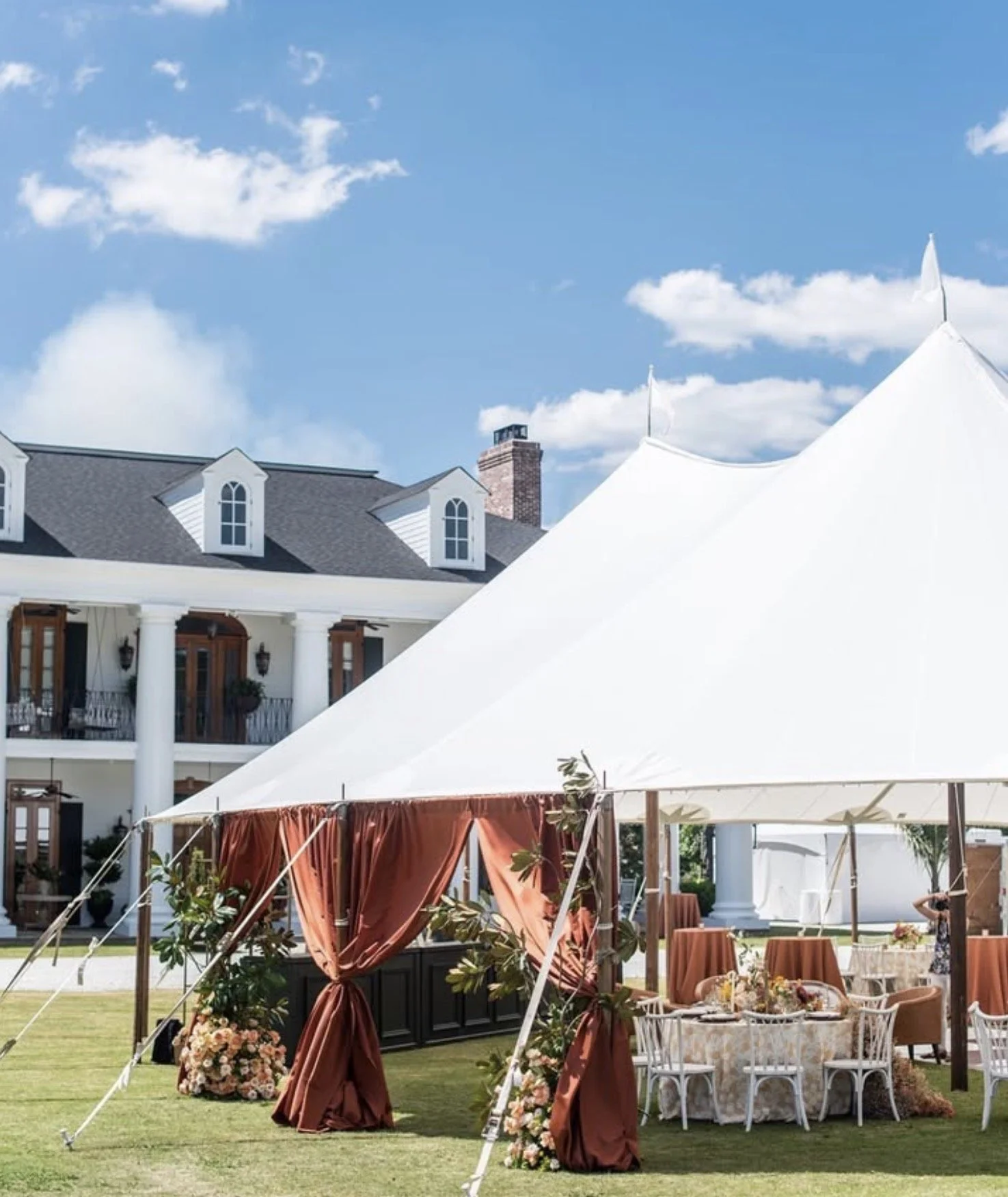 A white event tent set up outdoors on a lawn with a decorated table inside, surrounded by chairs, at a large house with white columns and a porch, under a blue sky with clouds.