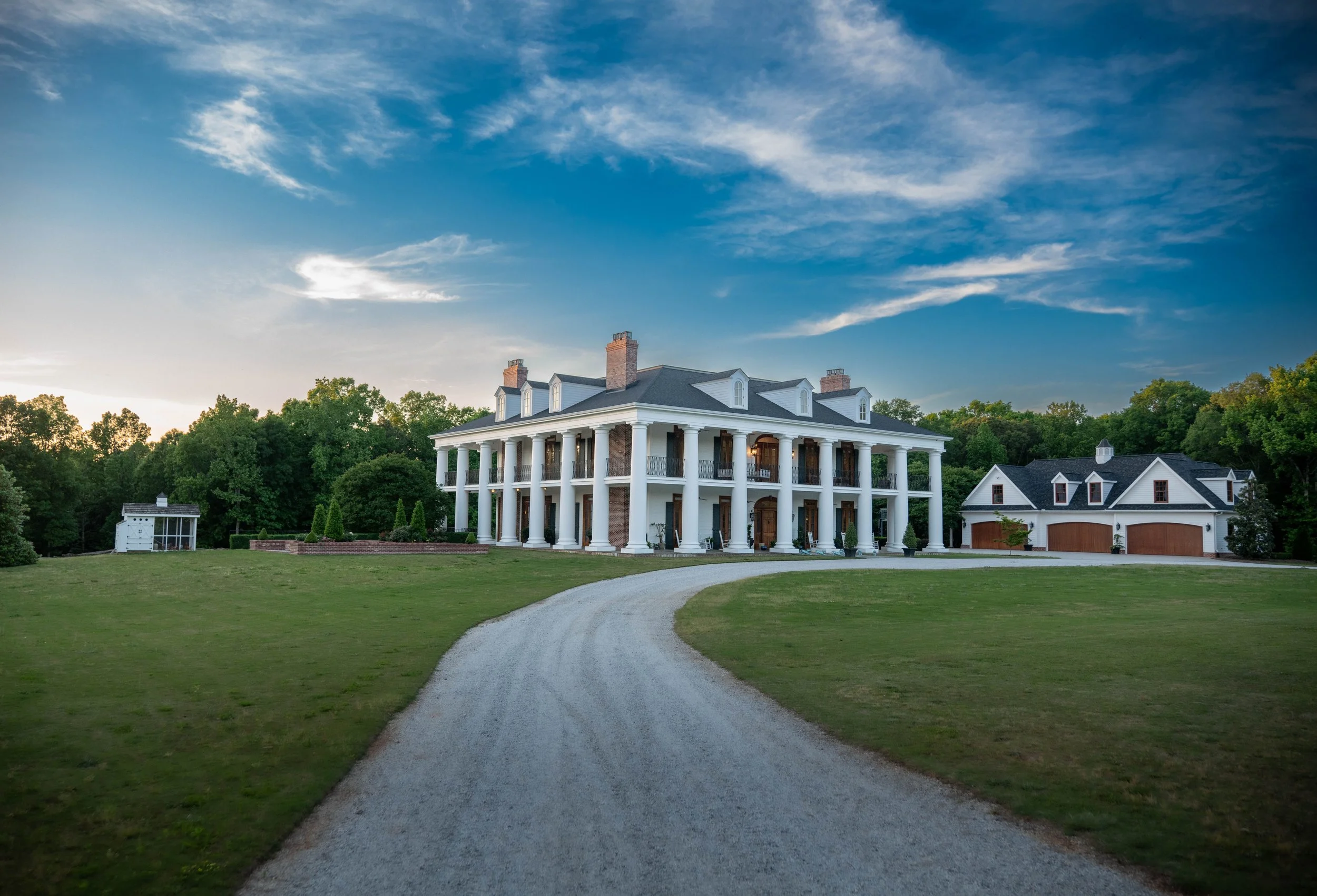 Large white mansion with tall columns in front, a circular gravel driveway, surrounded by green lawn and trees, with a blue sky overhead.