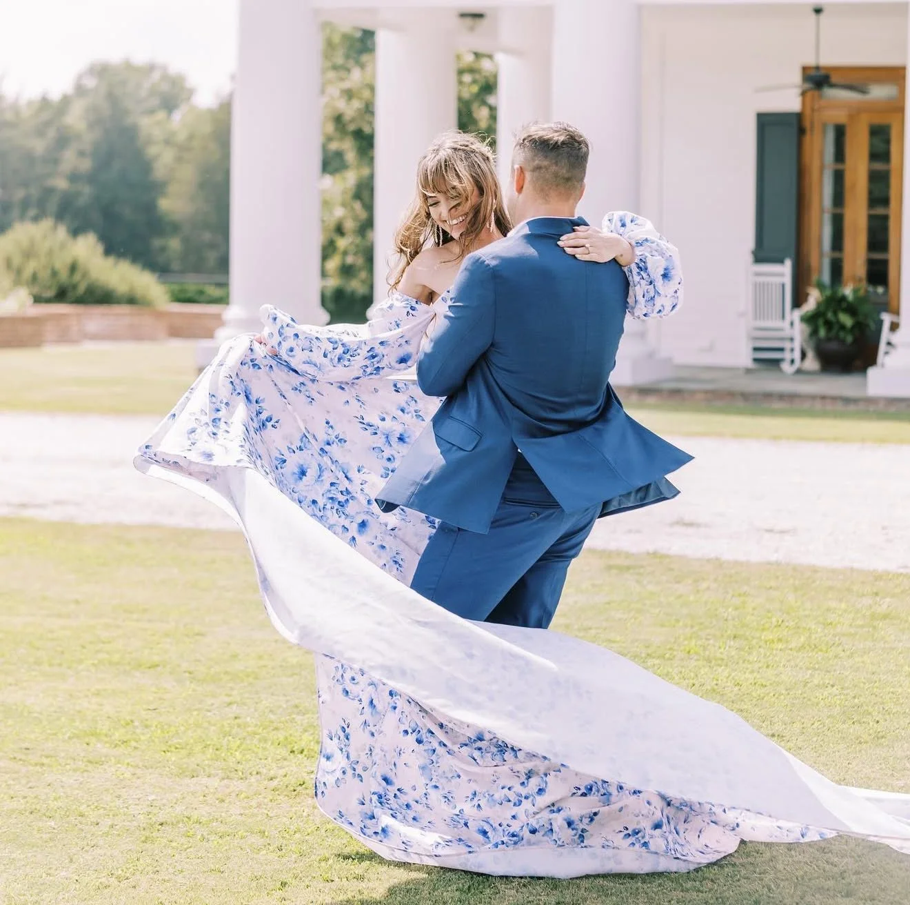 A man in a blue suit dancing with a woman in a floral dress outdoors in front of a white house.