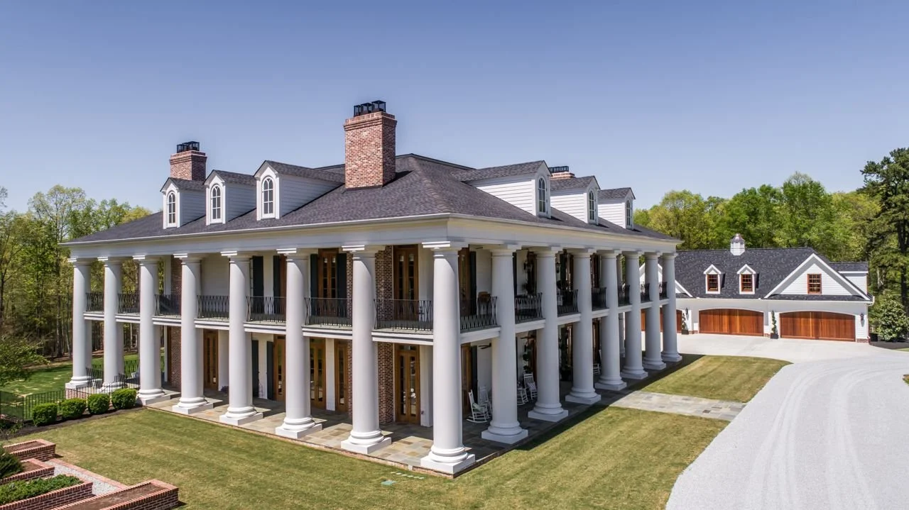 Large white mansion with tall columns, brick chimneys, and multiple dormer windows, surrounded by green trees and a well-maintained lawn.