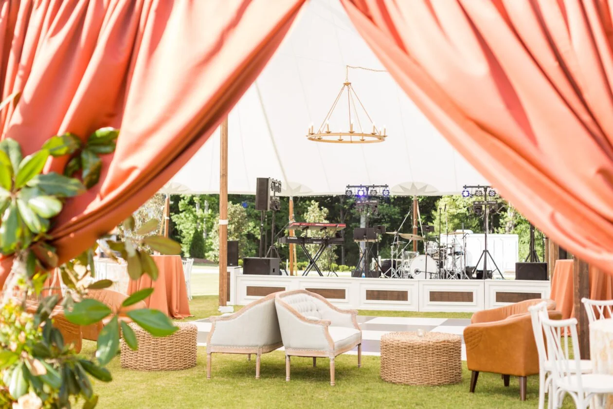 Outdoor event stage with musical instruments and equipment under a tent, surrounded by seating with pink curtains and greenery.