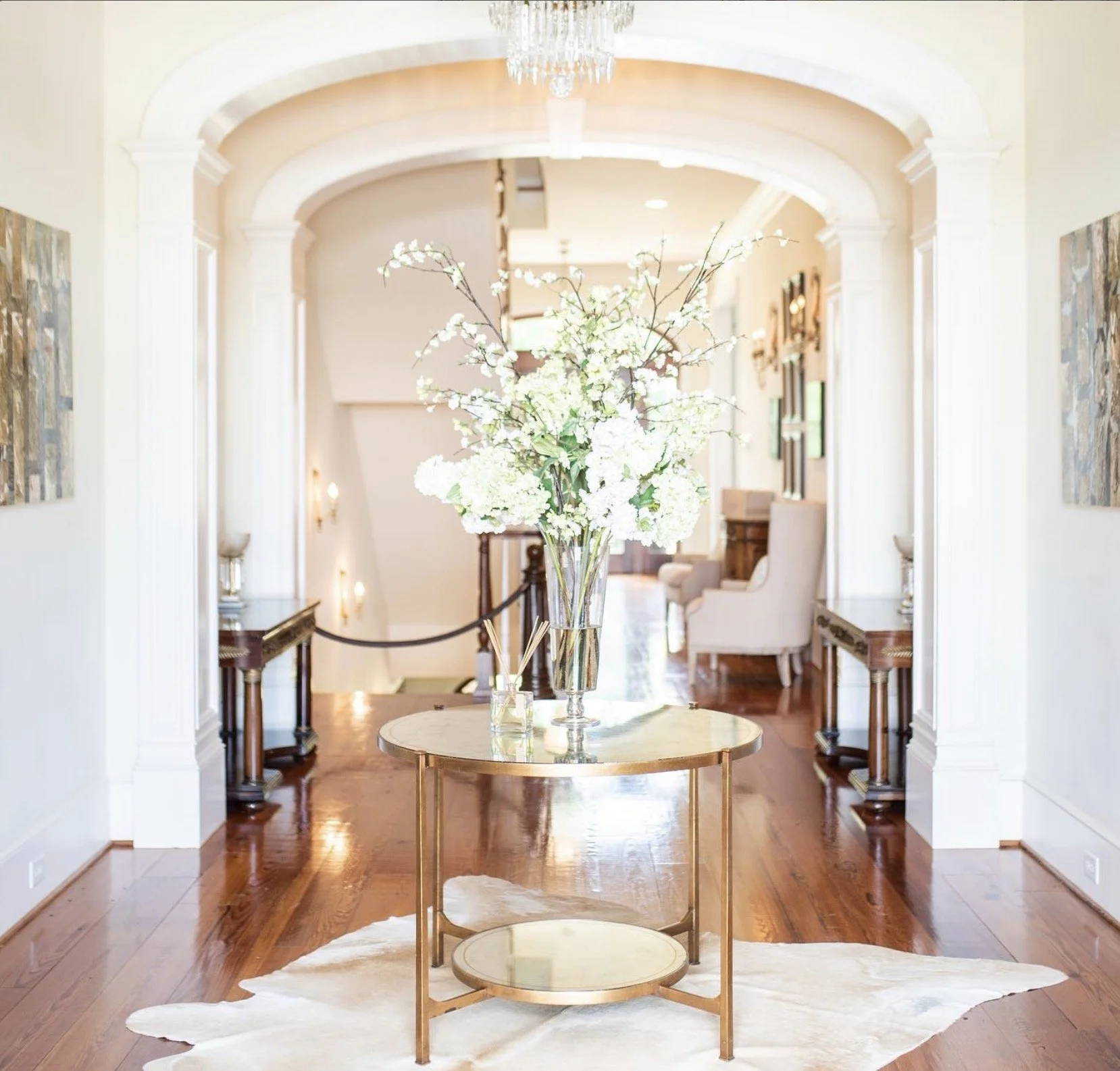 Elegant entryway with a gold round table holding a tall glass vase with white floral arrangement, wooden flooring, white walls, and framed artwork.