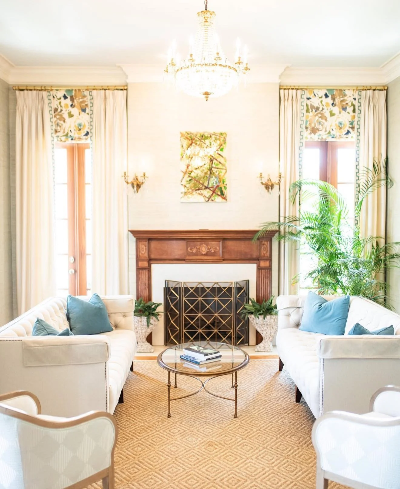 Elegant living room with two white sofas with blue throw pillows facing each other, a round glass coffee table with books, a fireplace with a wooden mantel, large windows with cream curtains and floral valances, a chandelier, a potted plant, and wall