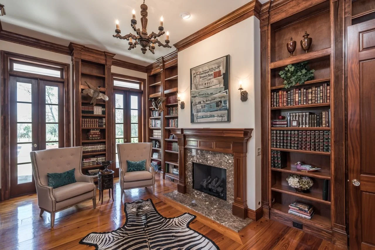 A cozy living room with wooden bookshelves, a marble fireplace, two beige armchairs with green pillows, a zebra print rug, and a chandelier.