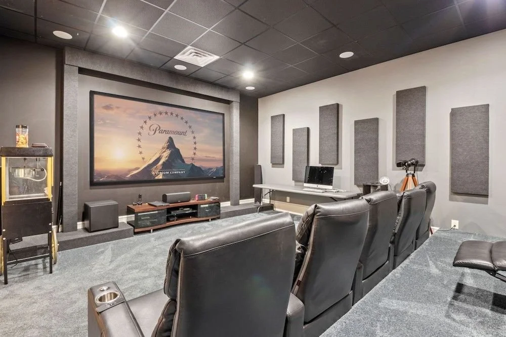 A home theater room with six leather recliner chairs facing a large screen displaying the Paramount logo with a mountain and sunset background. The room has gray walls with acoustic panels, a black ceiling, and a carpeted floor. There is a small tabl