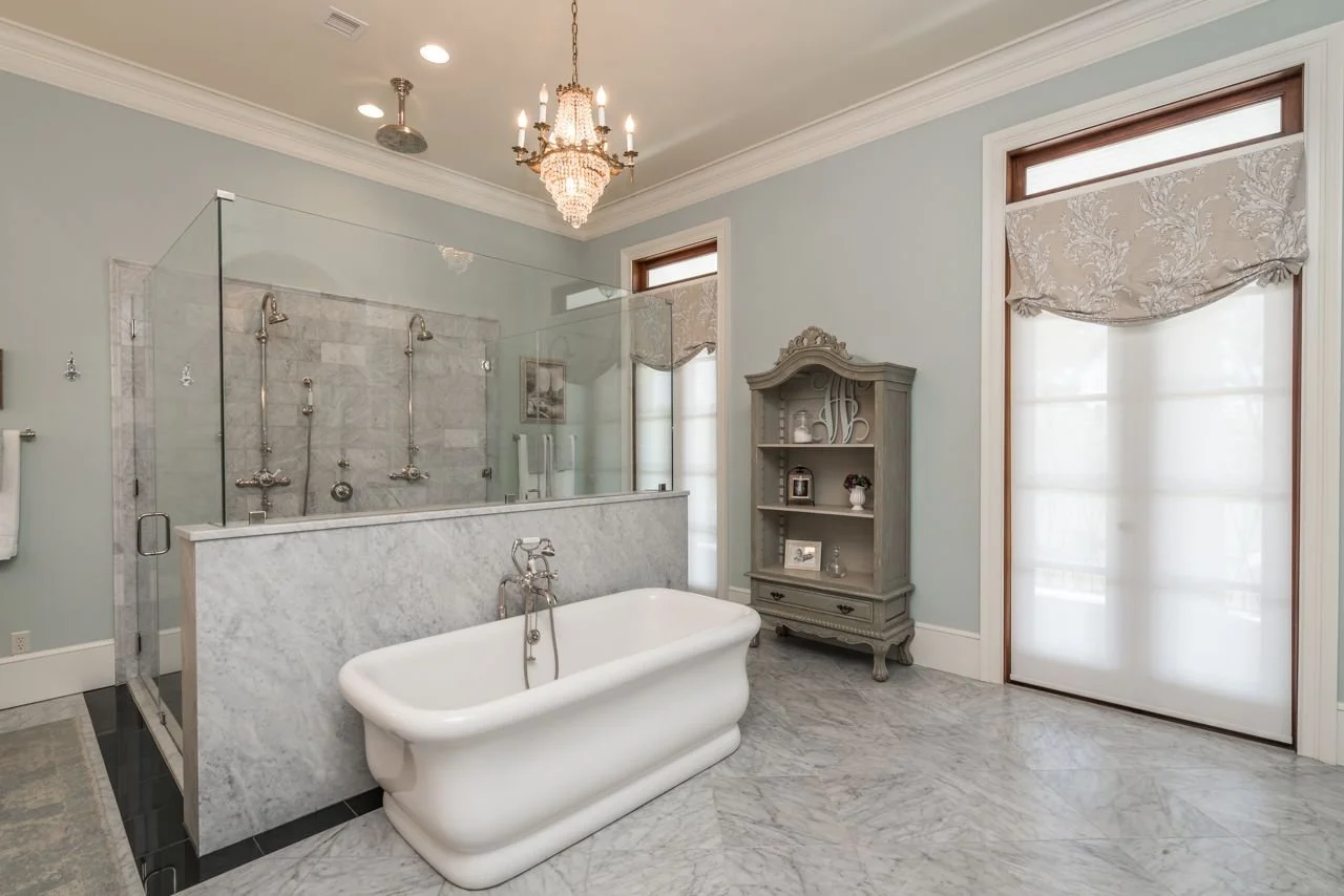Elegant bathroom with a clawfoot bathtub, marble flooring, and a glass-enclosed shower with marble accents. Two windows with patterned curtains, an ornate chandelier, and a decorative grey cabinet add to the luxurious decor.
