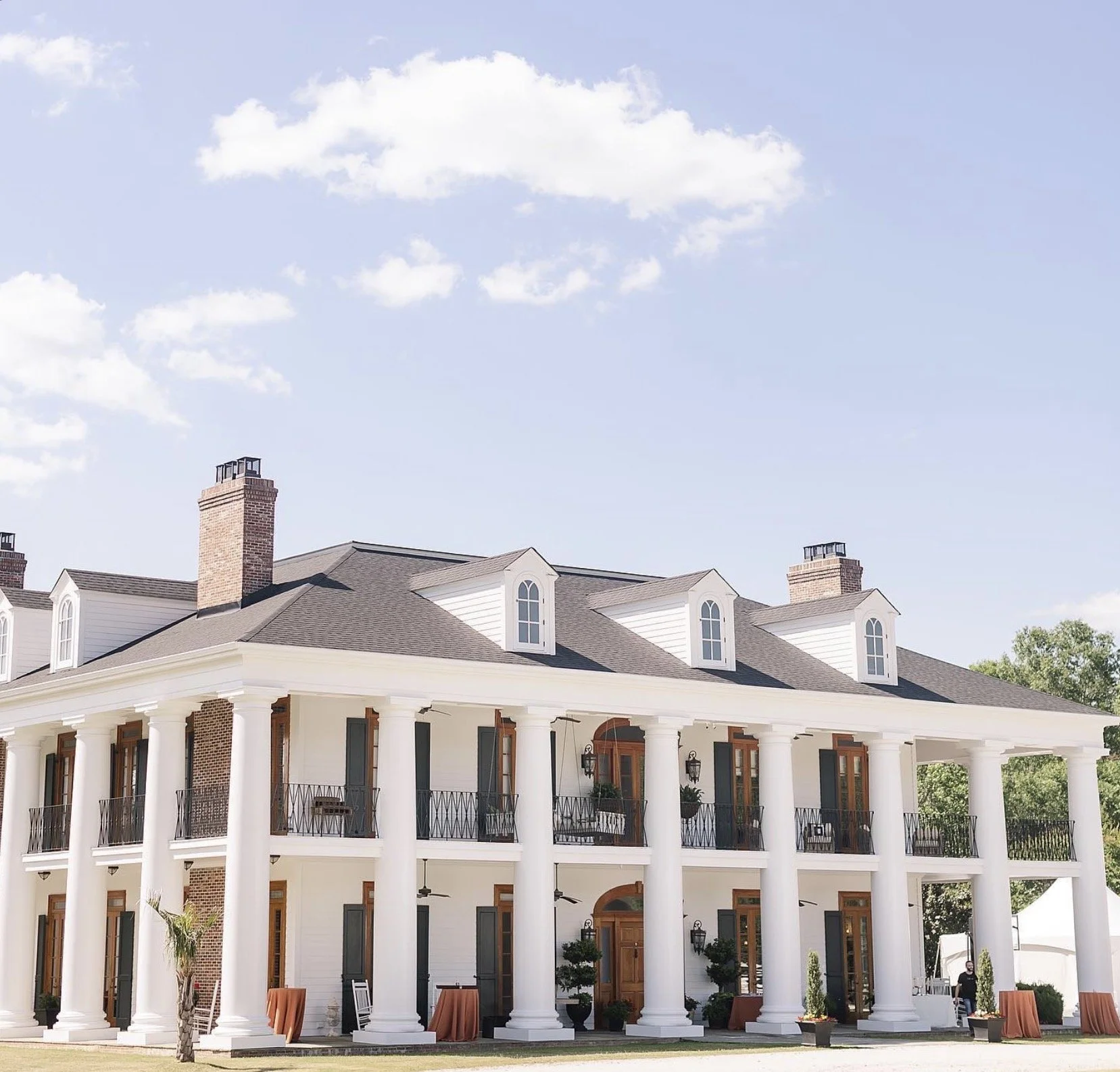 Large white mansion with tall columns, multiple balconies with black railings, and dormer windows on the roof; surrounded by greenery and under a partly cloudy sky.