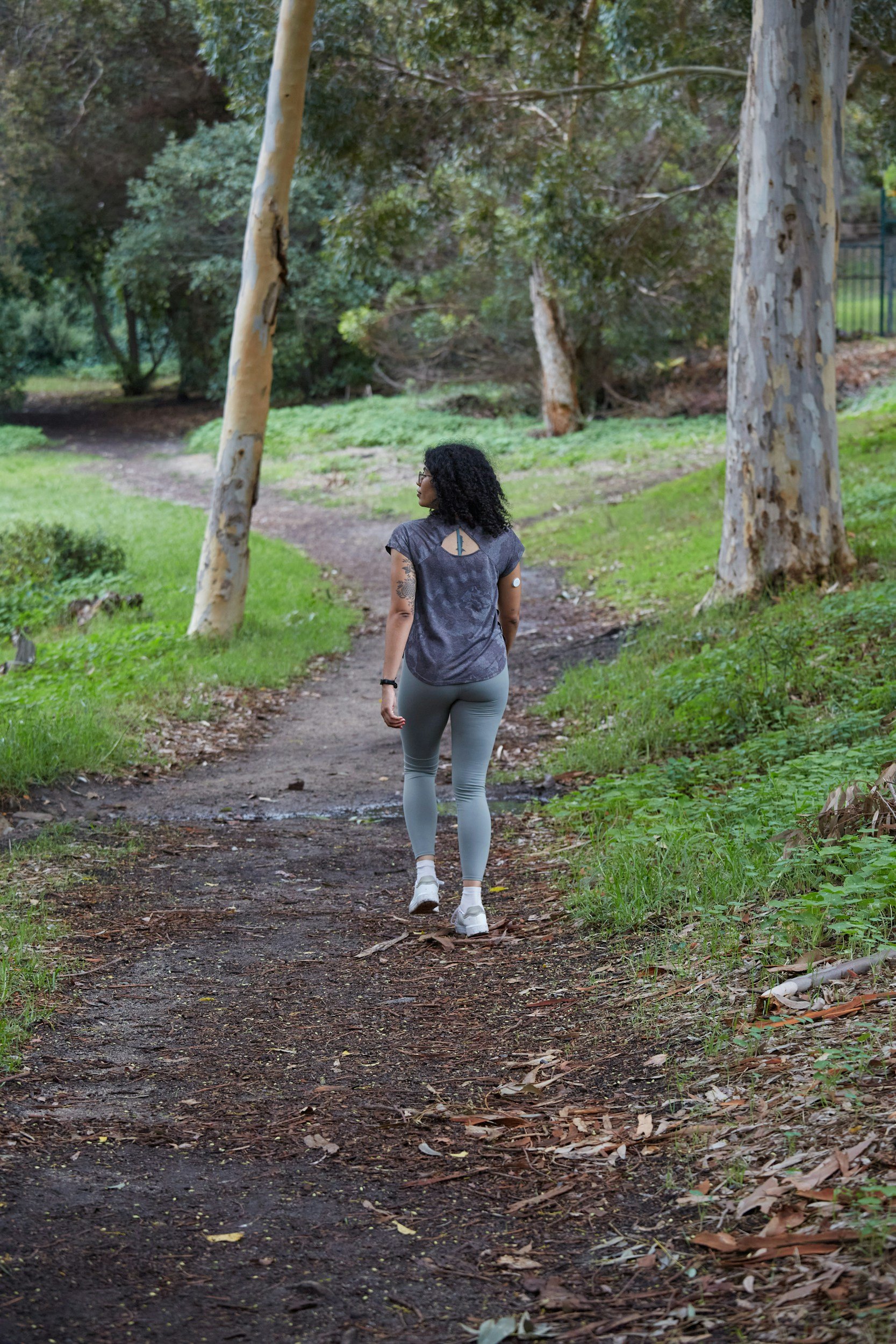 A person walking on a dirt trail in a wooded area during fall, wearing a black jacket, gray hoodie, black pants, and sneakers.