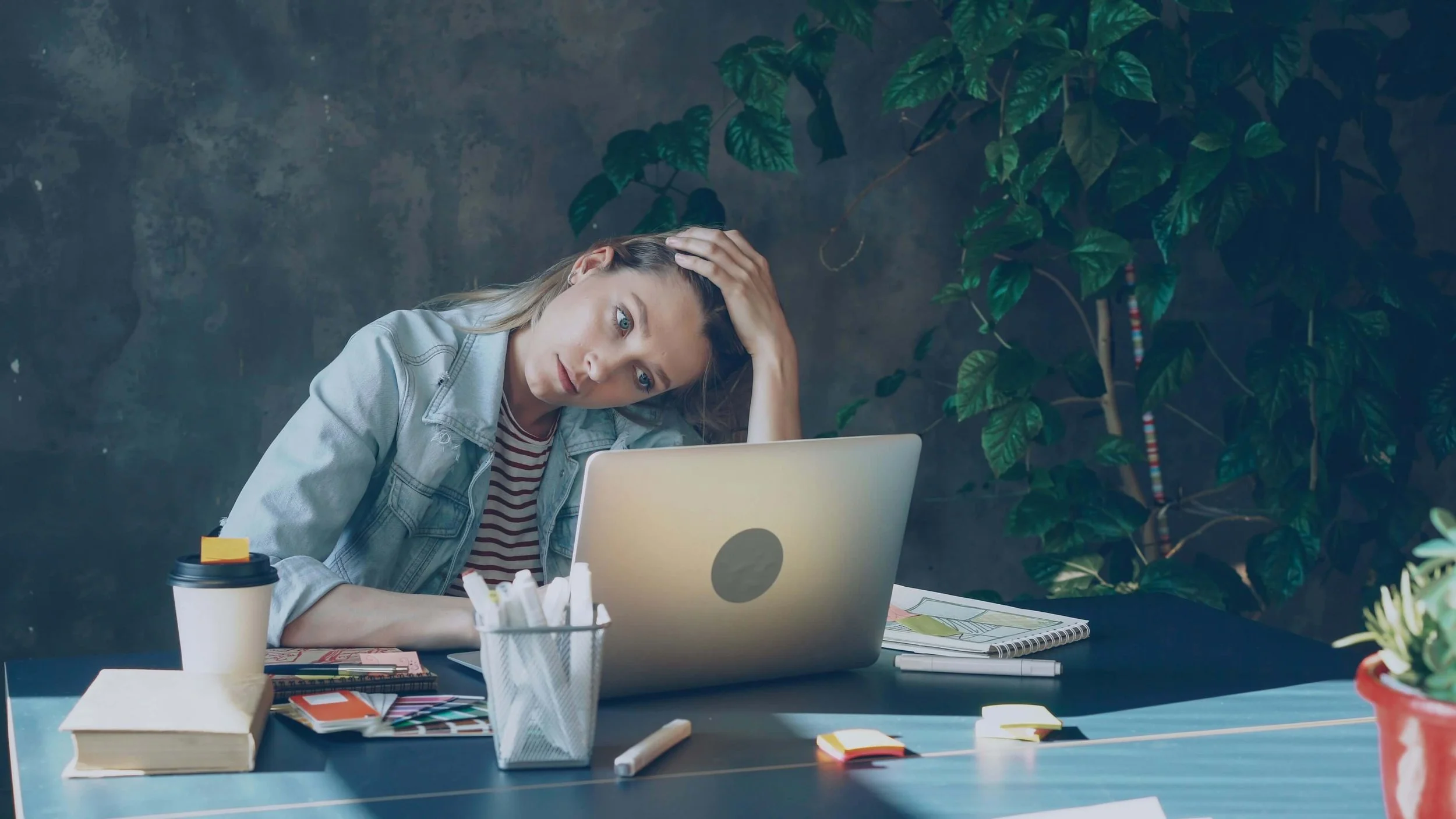 A woman sits at a desk with a laptop, holding her head with her hand, looking stressed or tired, surrounded by notebooks, pens, sticky notes, and a coffee cup.