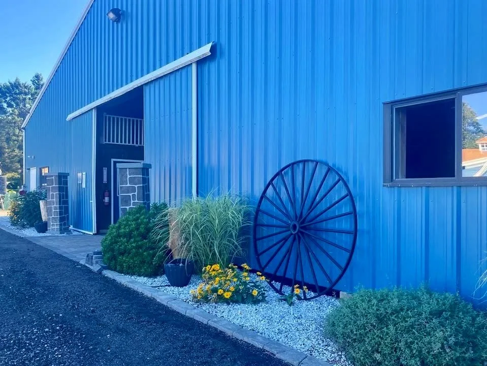 Blue metal building with a large wheel leaning against the wall, plants, flowers, and gravel landscaping outside