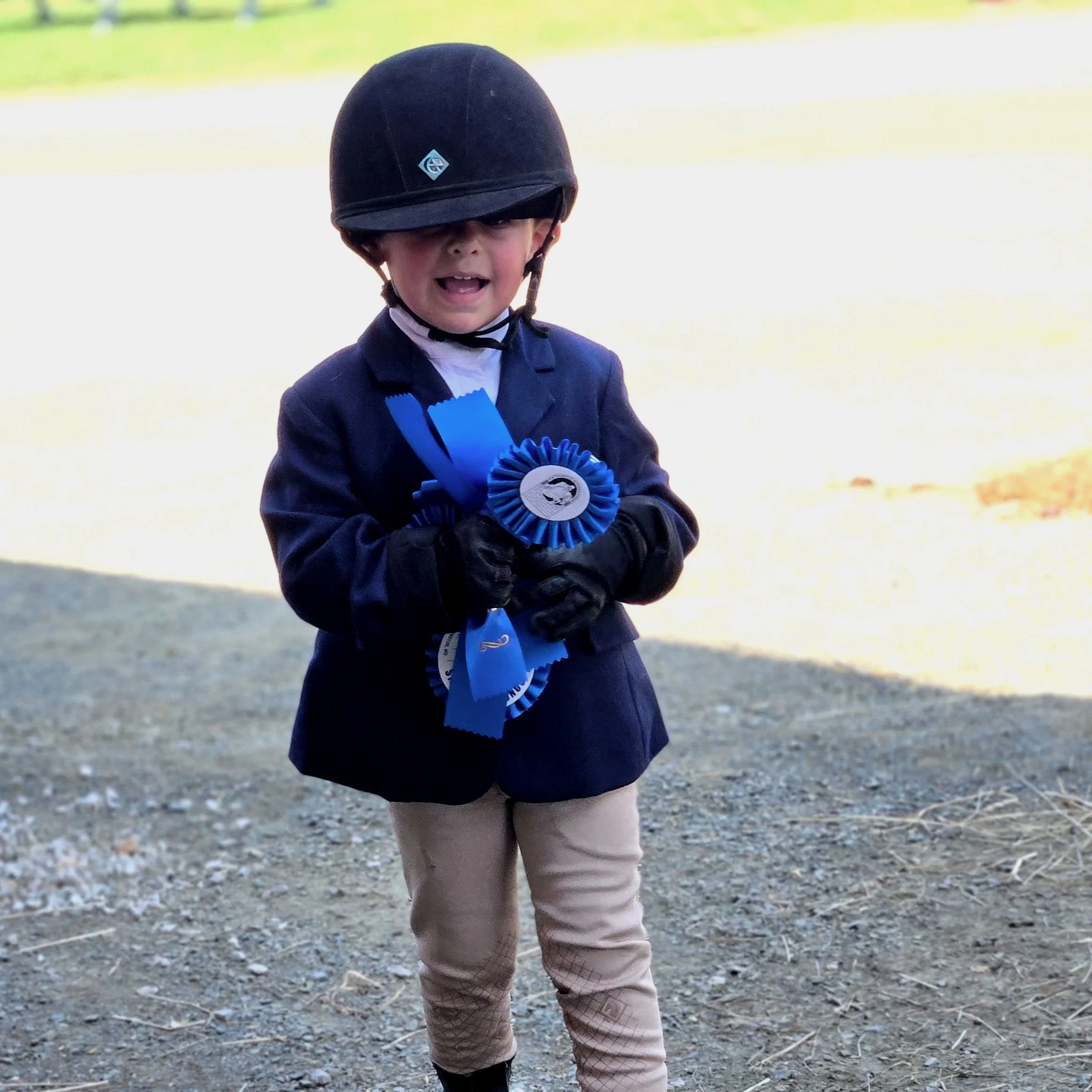 Young boy in equestrian attire, wearing a helmet, blazer, and riding pants, holding blue ribbons and medals, standing outdoors on a gravel surface.