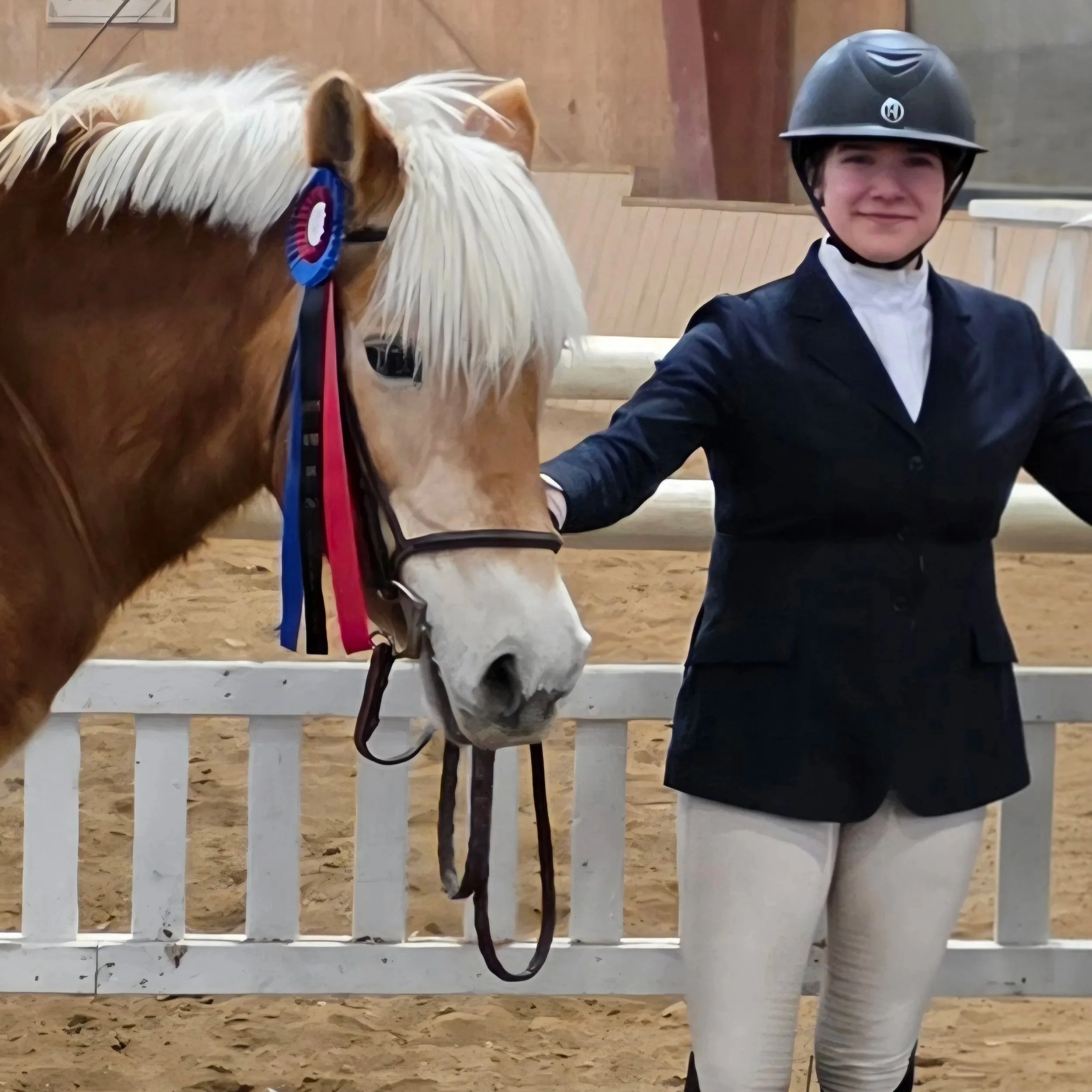 Person dressed in equestrian attire standing next to a palomino horse with a ribbon on the horse's bridle inside a riding arena.