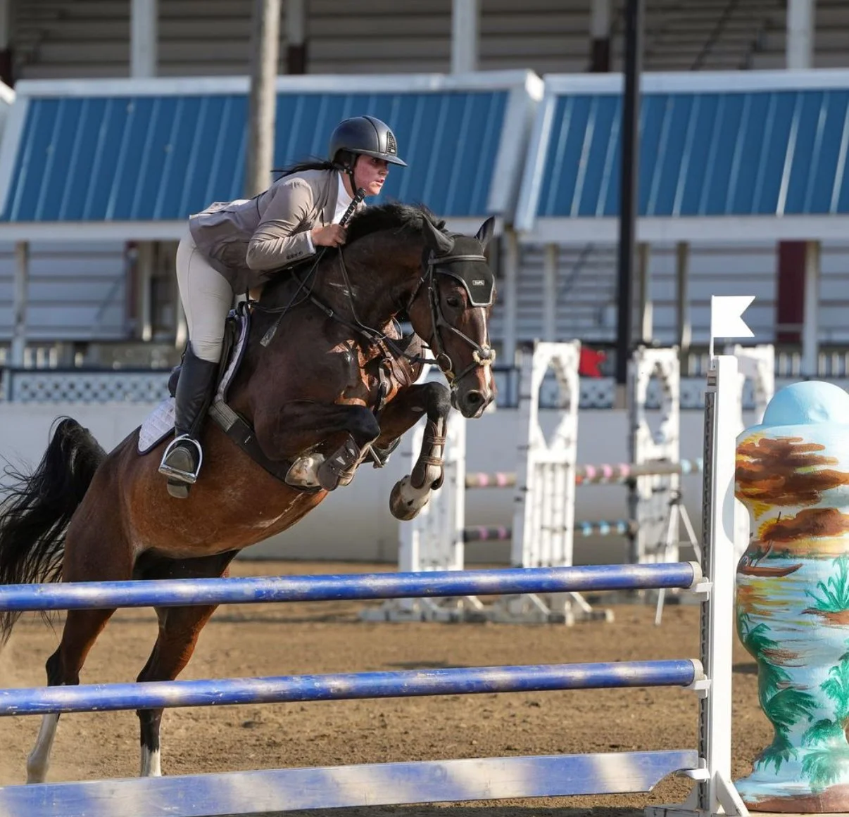 A woman wearing a helmet riding a brown horse during a show jumping event, with colorful decorations and barriers in the background.