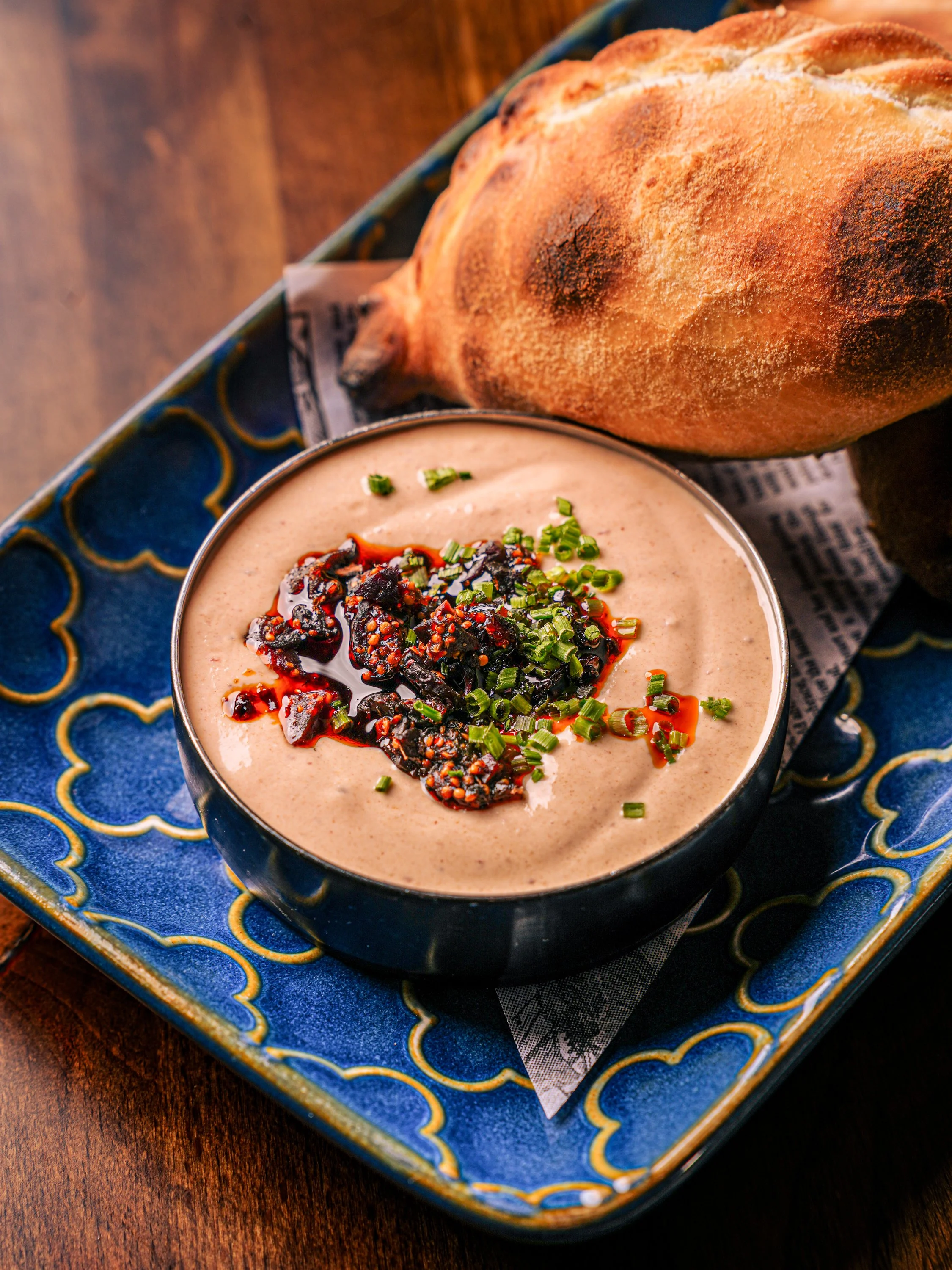 A bowl of creamy hummus topped with chopped green herbs and a red sauce, placed on a decorative blue and gold plate along with a toasted bread loaf on a wooden table.
