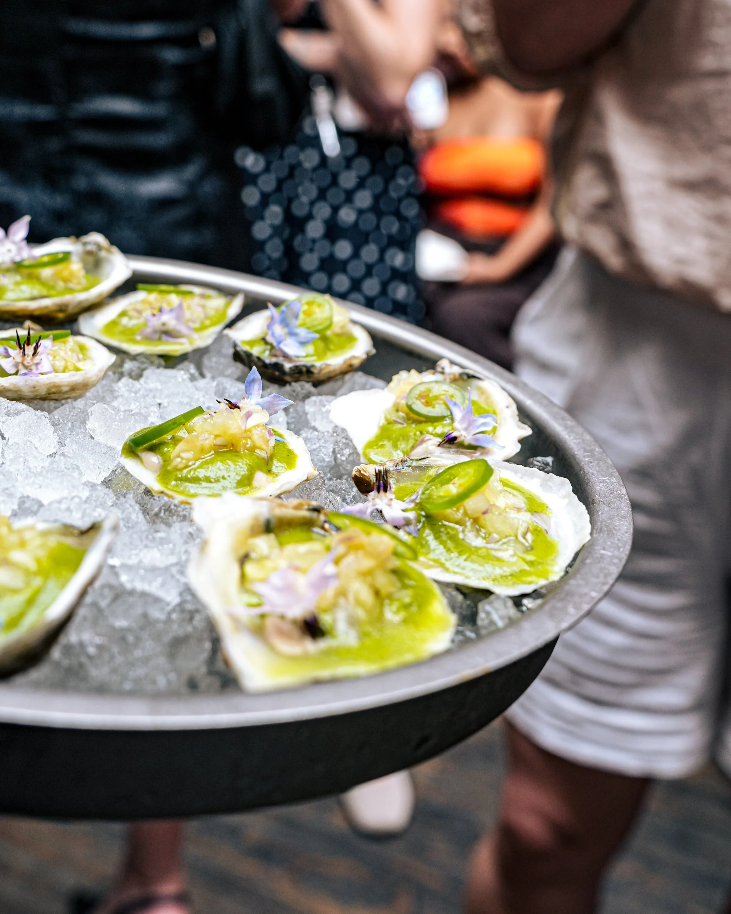 A tray of oysters topped with slices of lime, edible flowers, and a green sauce, served on ice at a seafood event.