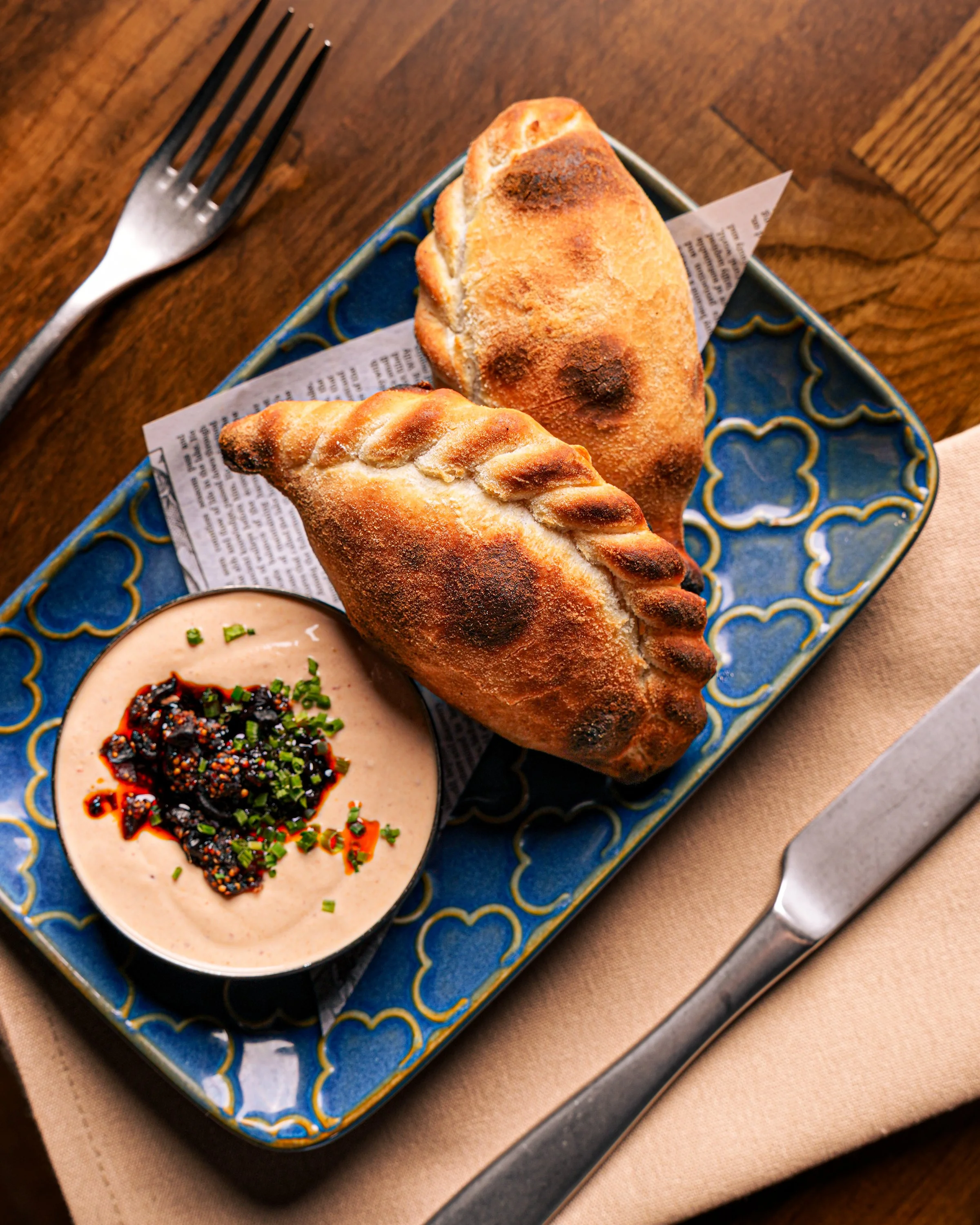 Two baked bread rolls on a decorative blue plate, with one roll having a twisted pattern and the other a smooth surface, accompanied by a bowl of creamy dipping sauce topped with chopped herbs and black sesame seeds, all on a wooden table.