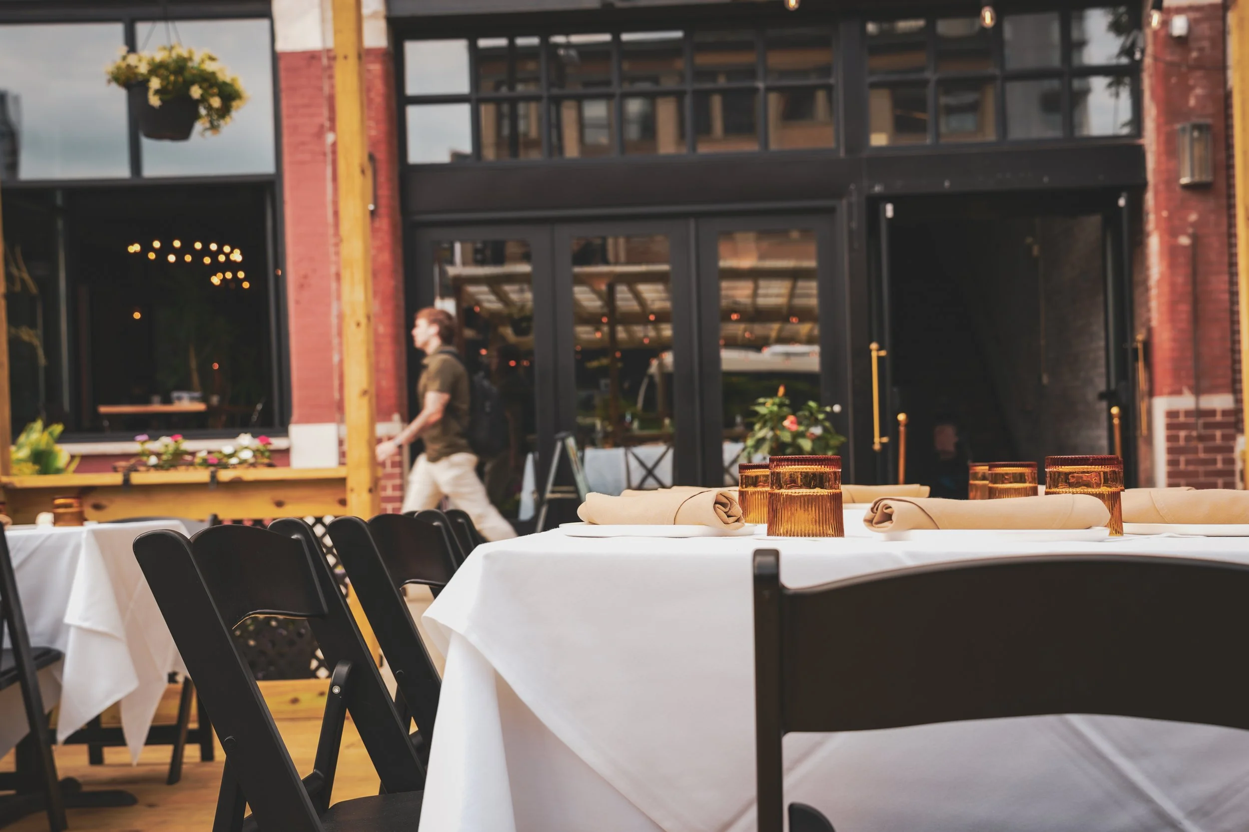 Indoor dining area with tables covered in white tablecloths, set with rolled napkins and amber-colored glasses, in front of large windows and brick walls, with a person walking past in the background.