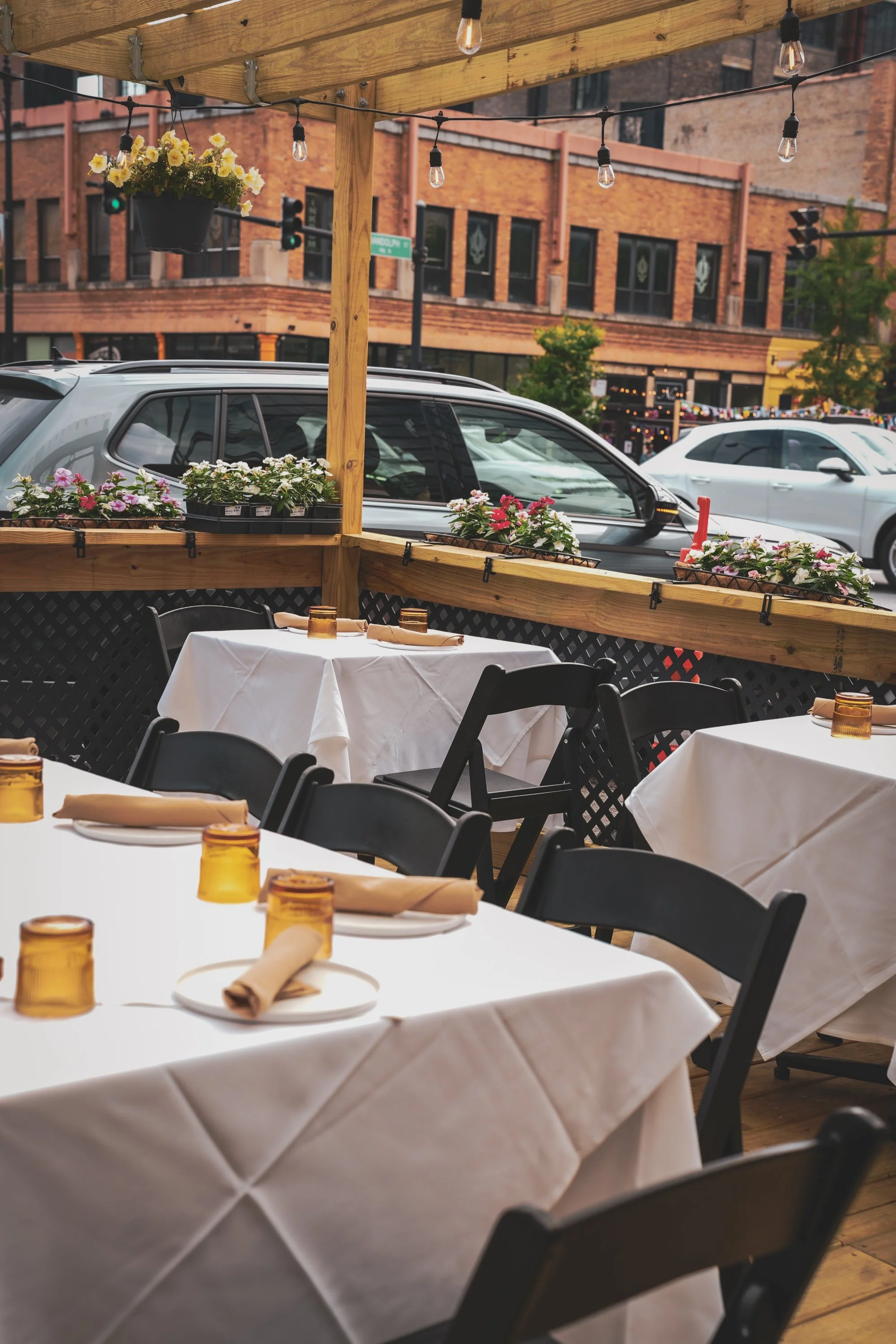 Outdoor dining area with tables covered in white tablecloths, napkins, amber glass cups, and black chairs. Potted flowers are on a wooden railing, with string lights hanging above. In the background, parked cars and buildings are visible.