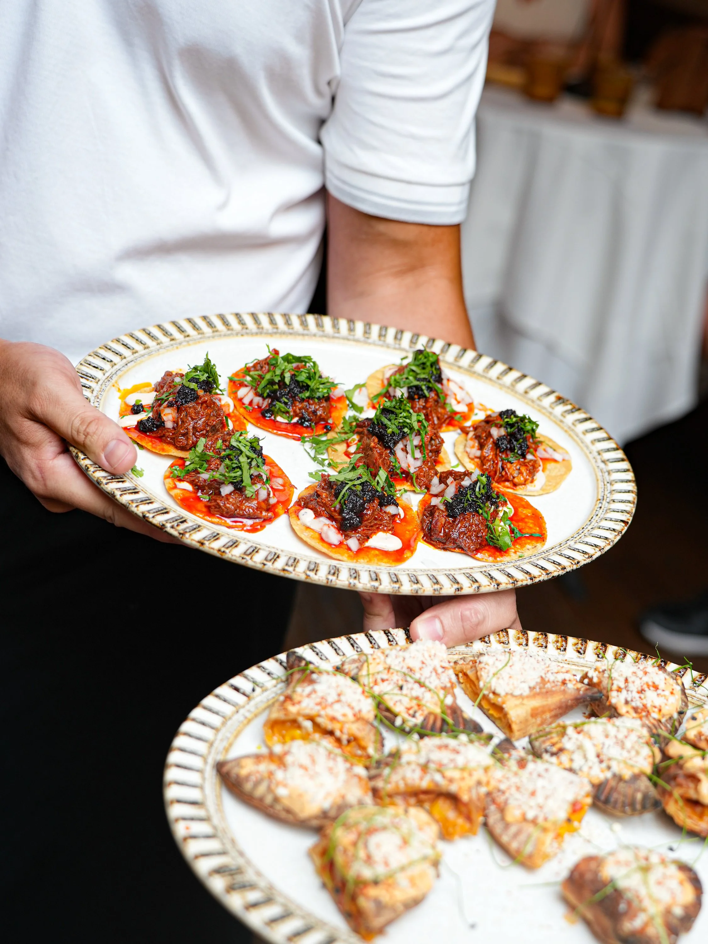 Person holding a plate of small bites with shredded meat, garnished with herbs and black sauce, on an ornate white plate with a gold and black border.