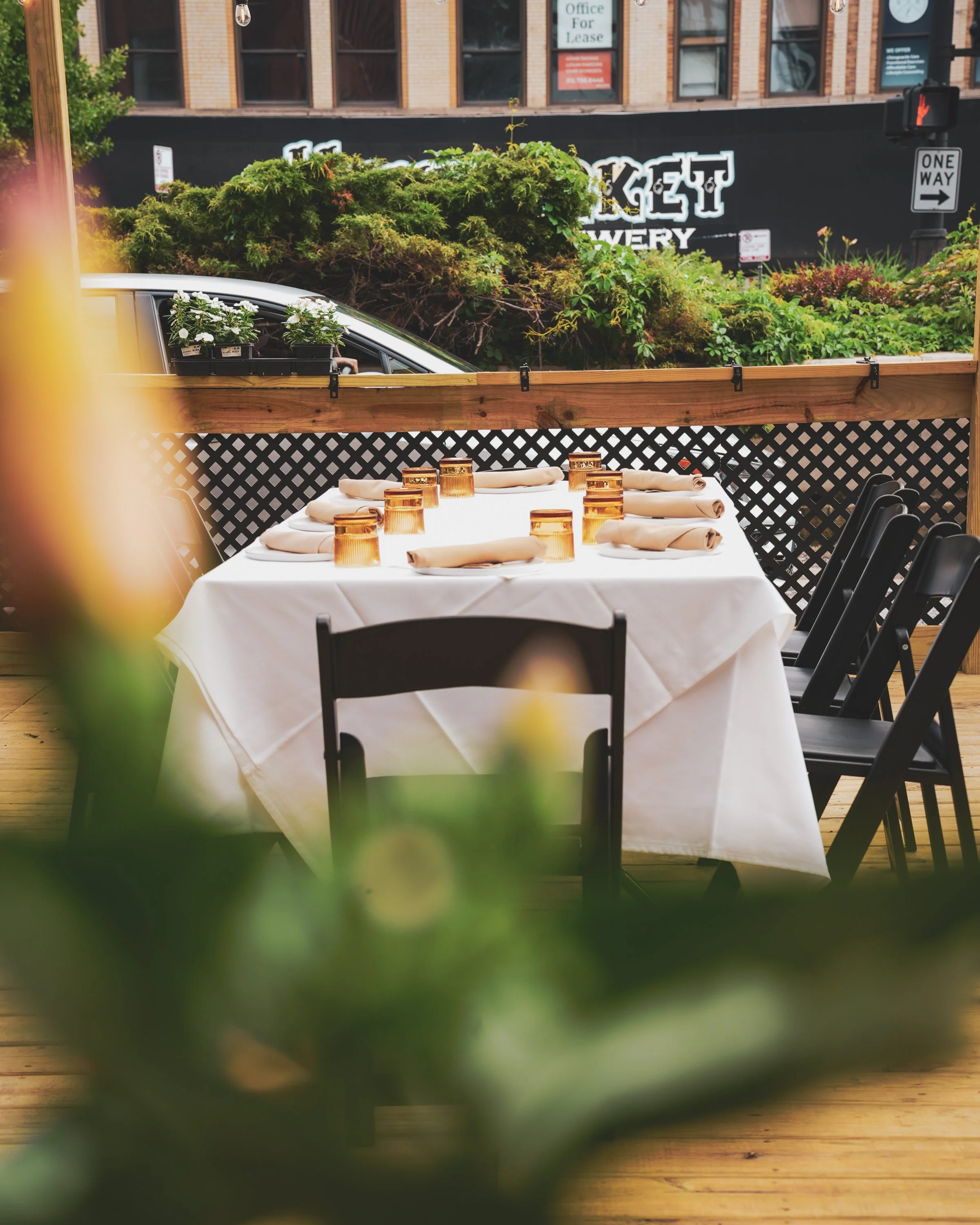 A table set for dining with a white tablecloth, beige napkins, and amber-colored glasses, positioned on a wooden deck, with lush green plants and bushes in the background, and a building with signs and a traffic light visible beyond.