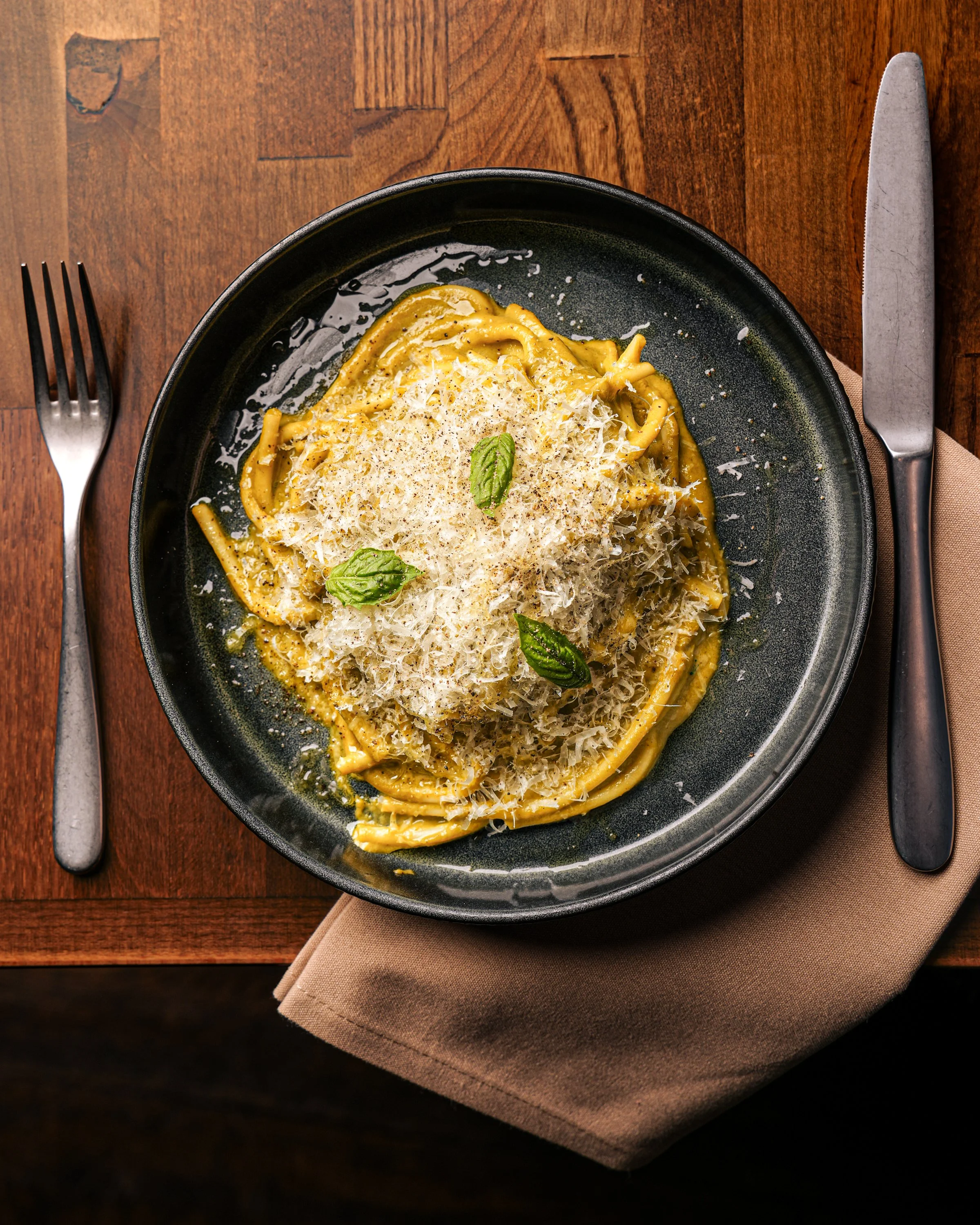 A black plate of spaghetti with a creamy sauce, garnished with grated cheese and fresh basil leaves, on a wooden table with a fork on the left and a knife on the right.