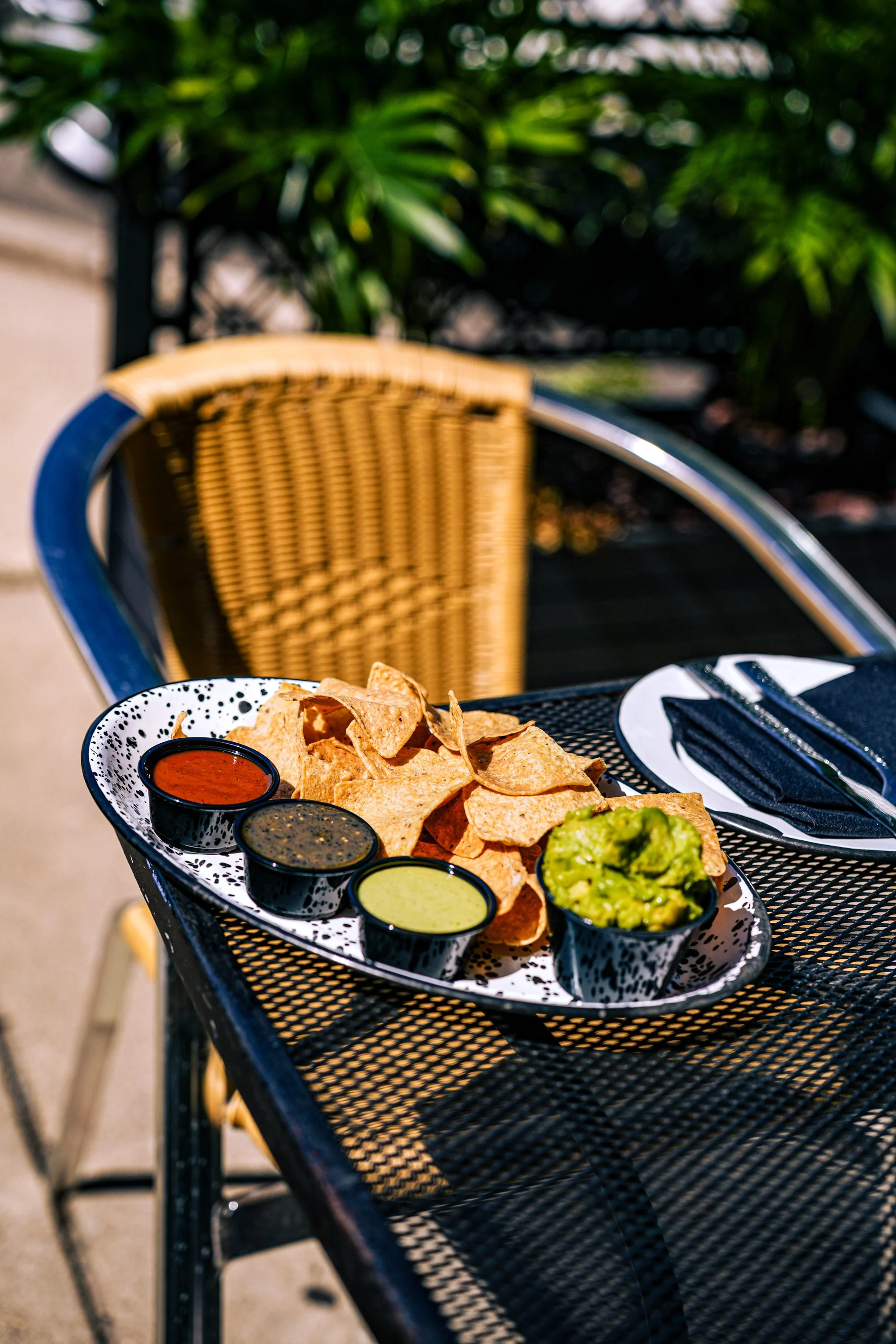 A plate of nachos with three small bowls of salsa, guacamole, and creamy sauce on a black metal outdoor table with a black napkin, fork, and knife. In the background, a rattan chair and green foliage are visible.