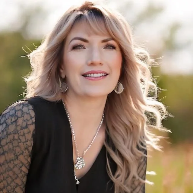 A photo of Jessica, death doula with blonde, wavy hair, wearing a black dress with lace sleeves, jewelry including earrings and a necklace, smiling outdoors with a blurred green background.