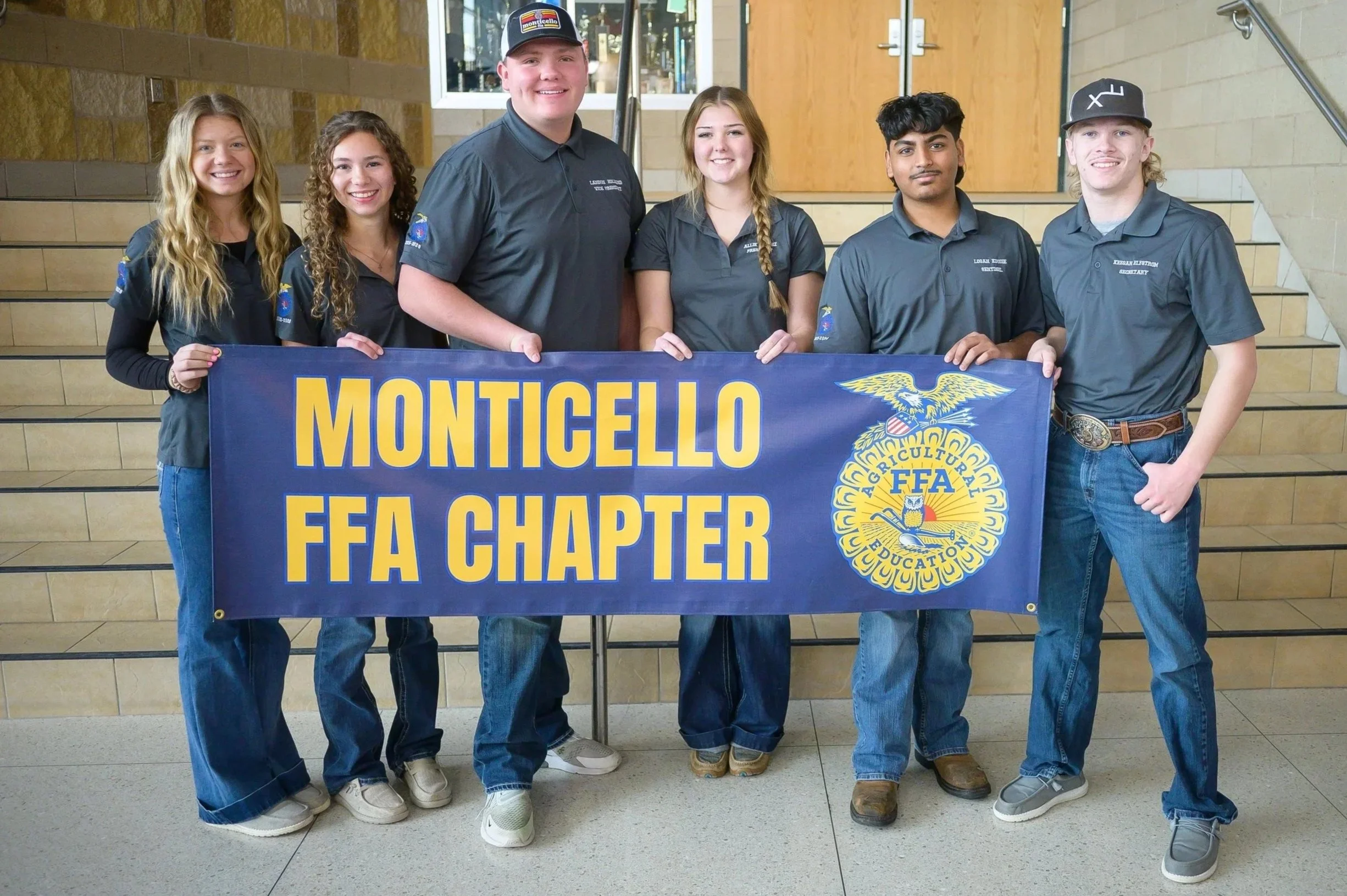 Group of six students holding a blue banner that reads 'Monticello FFA Chapter' with the FFA emblem in front of a staircase in a school hallway.
