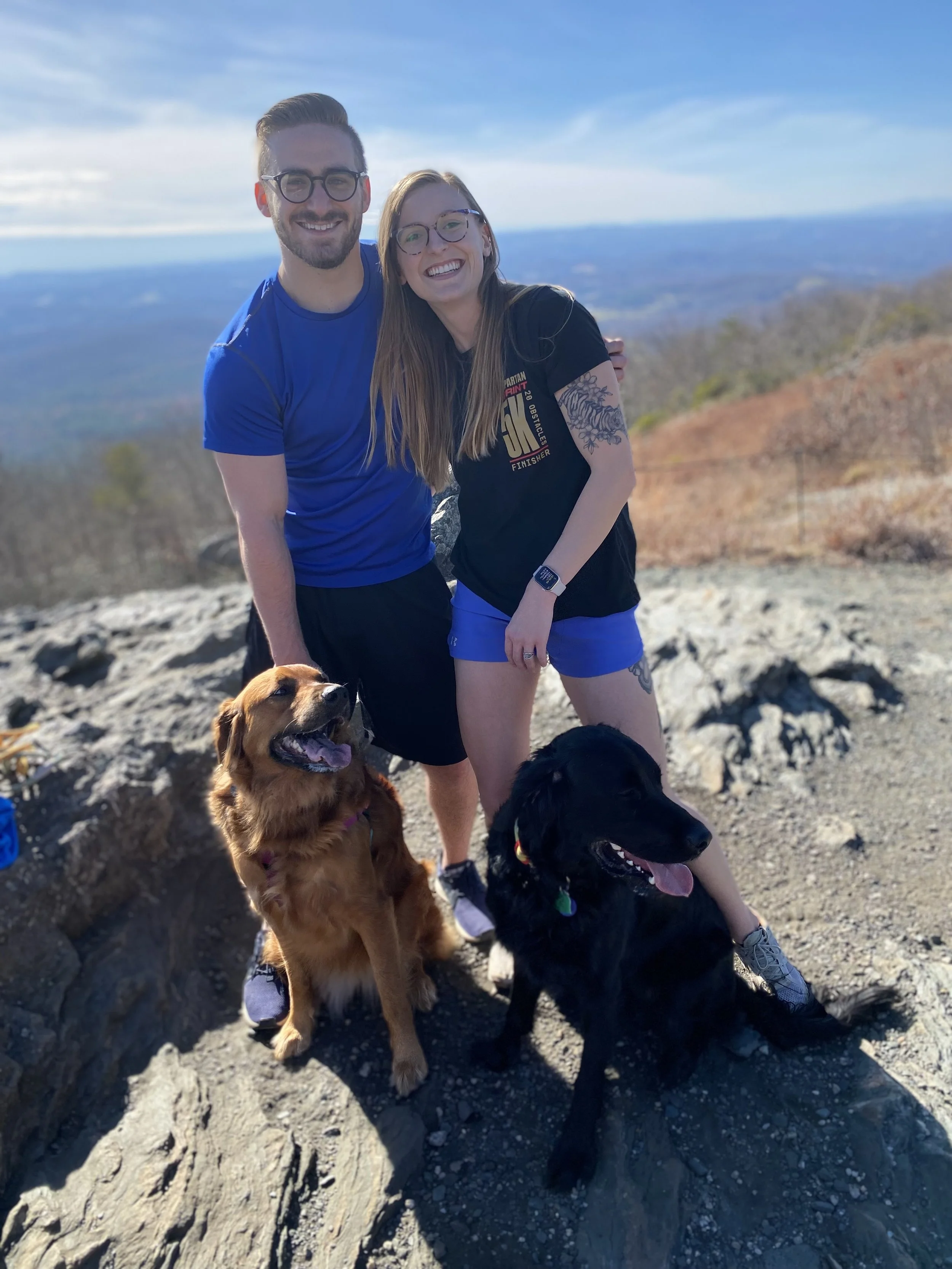 A smiling man and woman with glasses, standing outdoors on a rocky hilltop with two dogs, a golden retriever and a black retriever, during daytime with a view of distant mountains and a blue sky with some clouds.