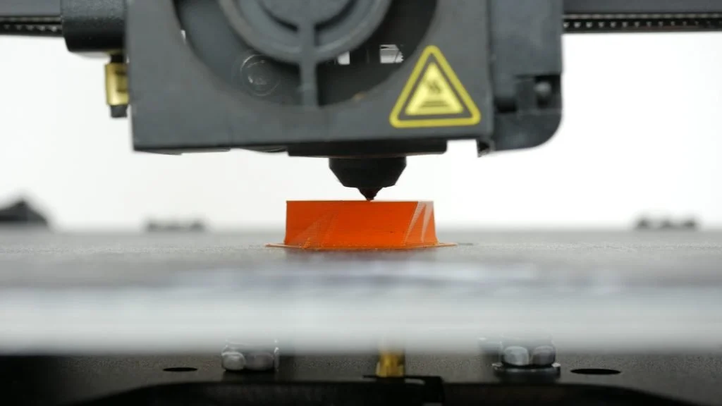 Close-up of a 3D printer nozzle printing an orange, circular object on the print bed.