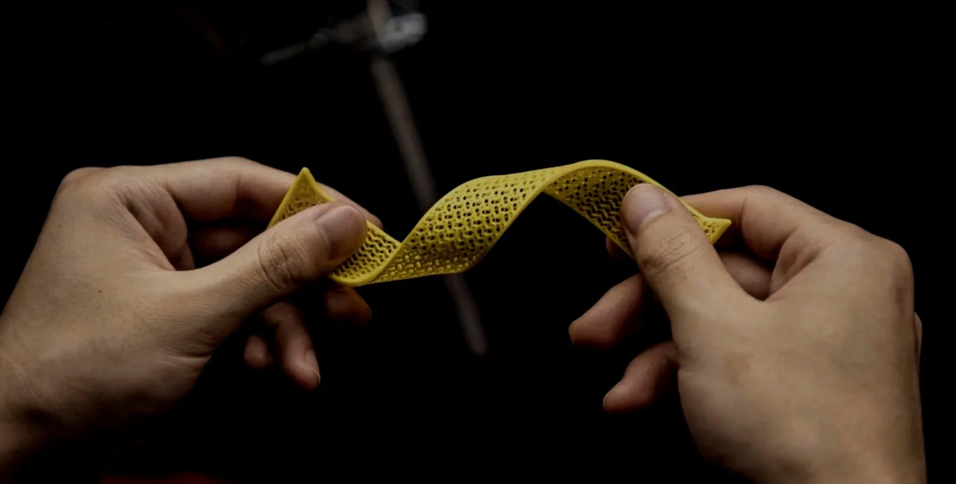 Hands holding a flexible, yellow, perforated strip of material against a dark background.