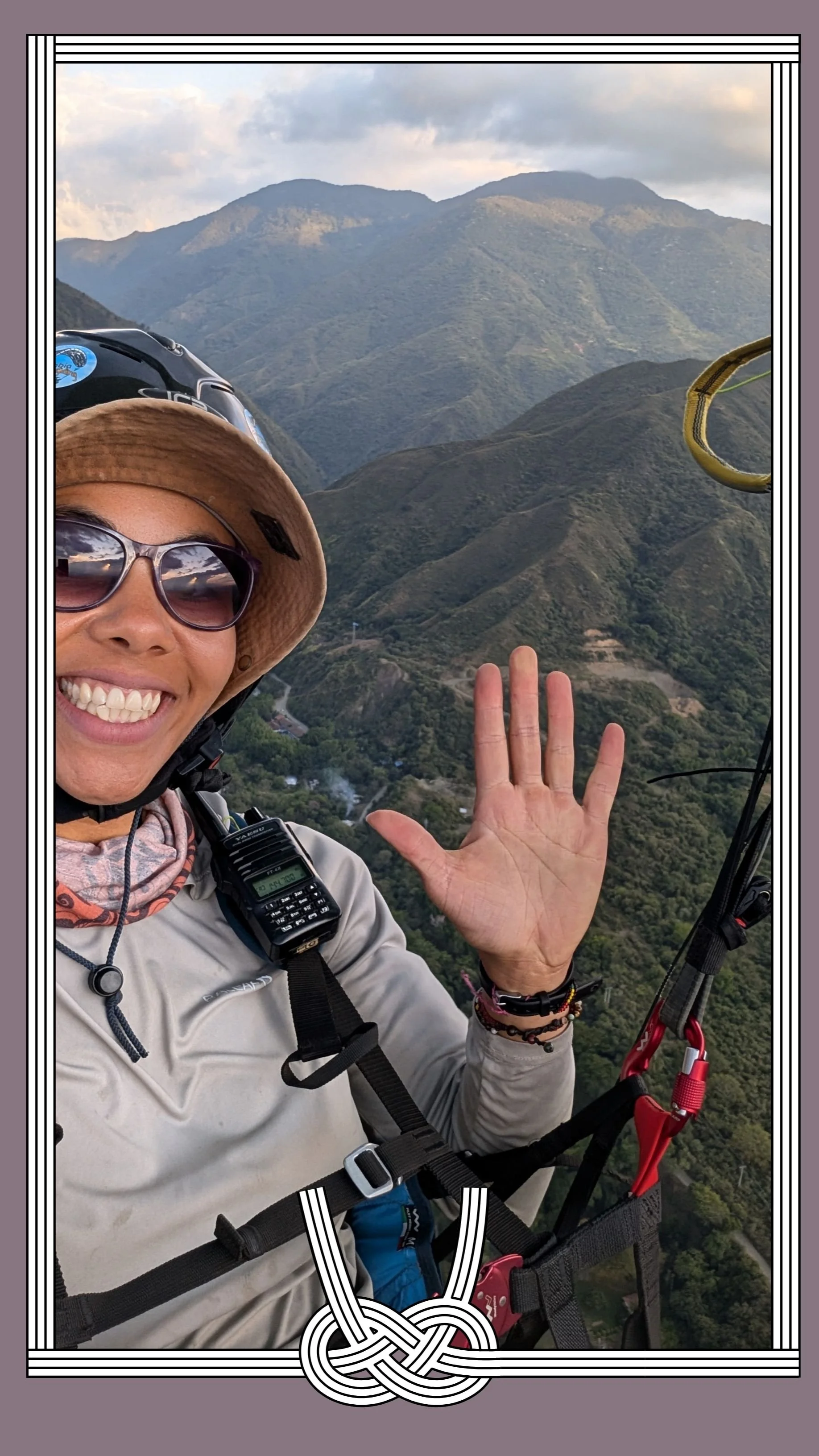 A smiling woman in outdoor gear, wearing sunglasses and a wide-brimmed hat, taking a selfie while paragliding over green mountainous terrain in the background.